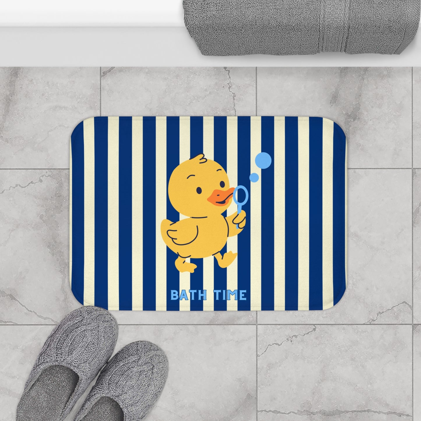A rectangular bath mat with rounded corners featuring navy blue and cream vertical stripes, a yellow cartoon duck blowing blue bubbles, and the words 'BATH TIME' in light blue. The mat is shown on a light gray marble tile floor with gray slippers and a folded gray towel nearby.