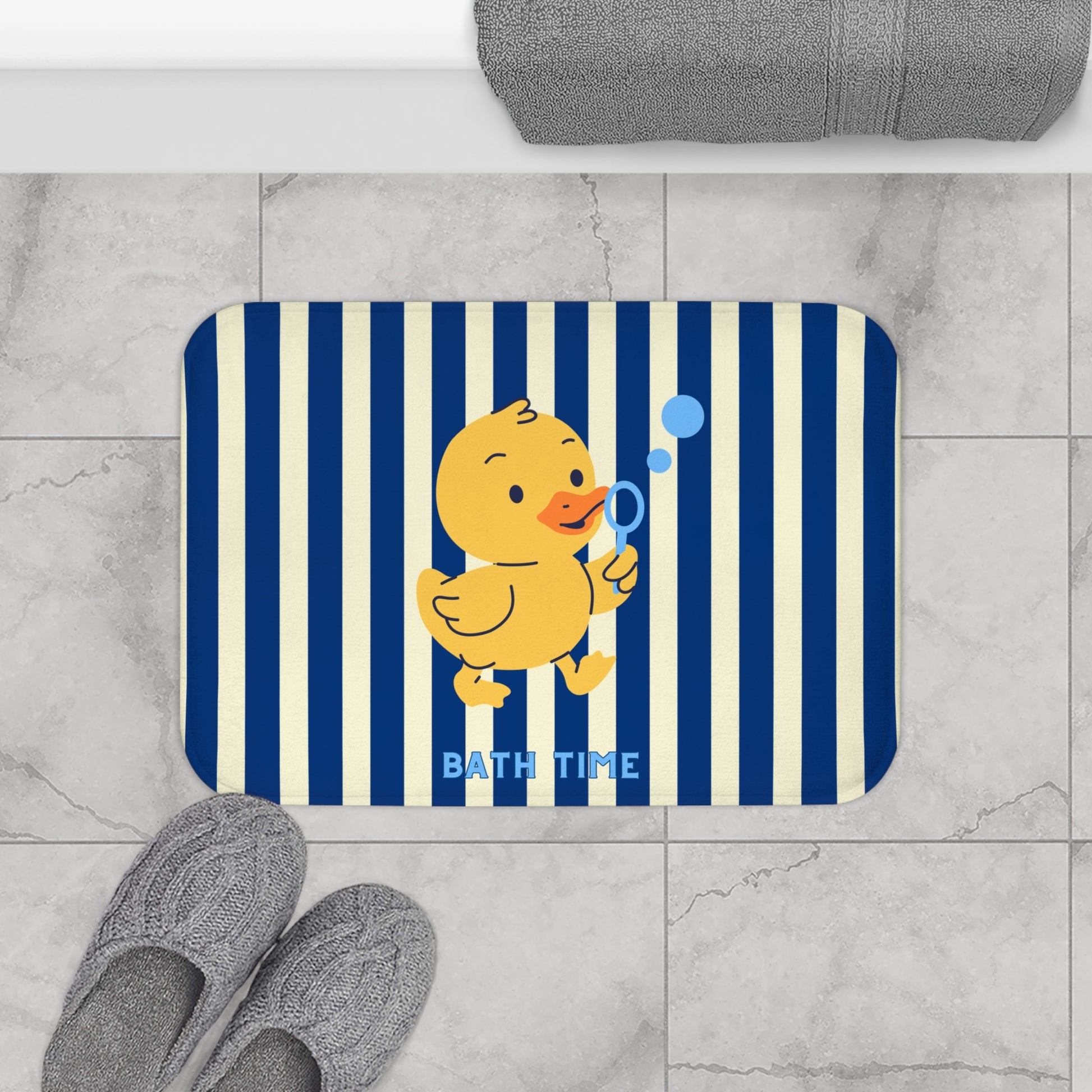 A rectangular bath mat with rounded corners featuring navy blue and cream vertical stripes, a yellow cartoon duck blowing blue bubbles, and the words 'BATH TIME' in light blue. The mat is shown on a light gray marble tile floor with gray slippers and a folded gray towel nearby.