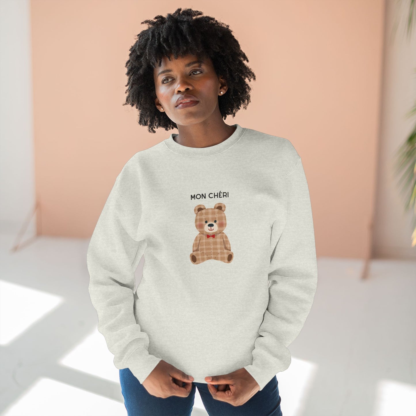 Person wearing a light gray sweatshirt with a teddy bear design and text, standing against a peach-colored wall.
