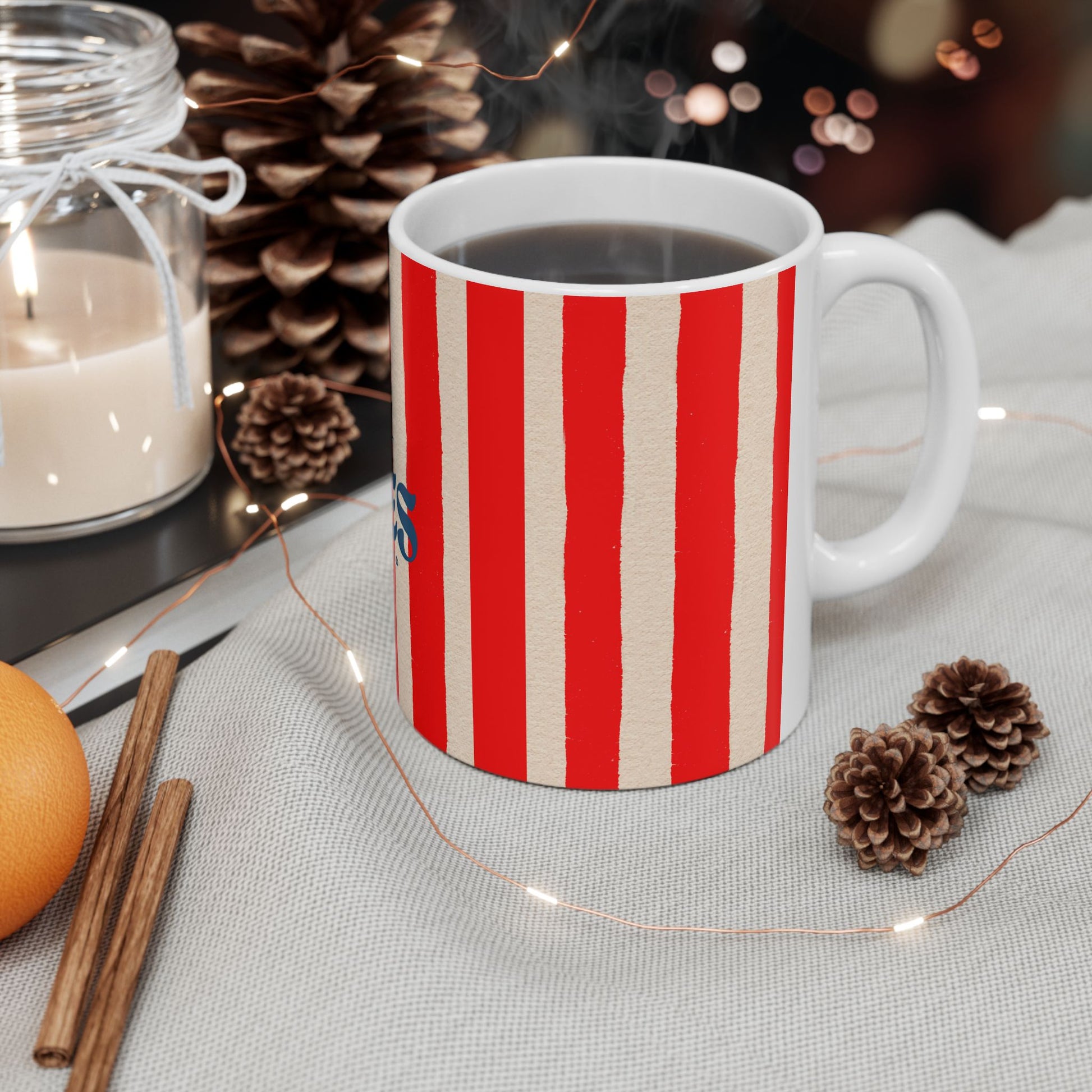 Red and white striped mug with a Christmas theme on a table with pinecones, an orange, and cinnamon sticks.