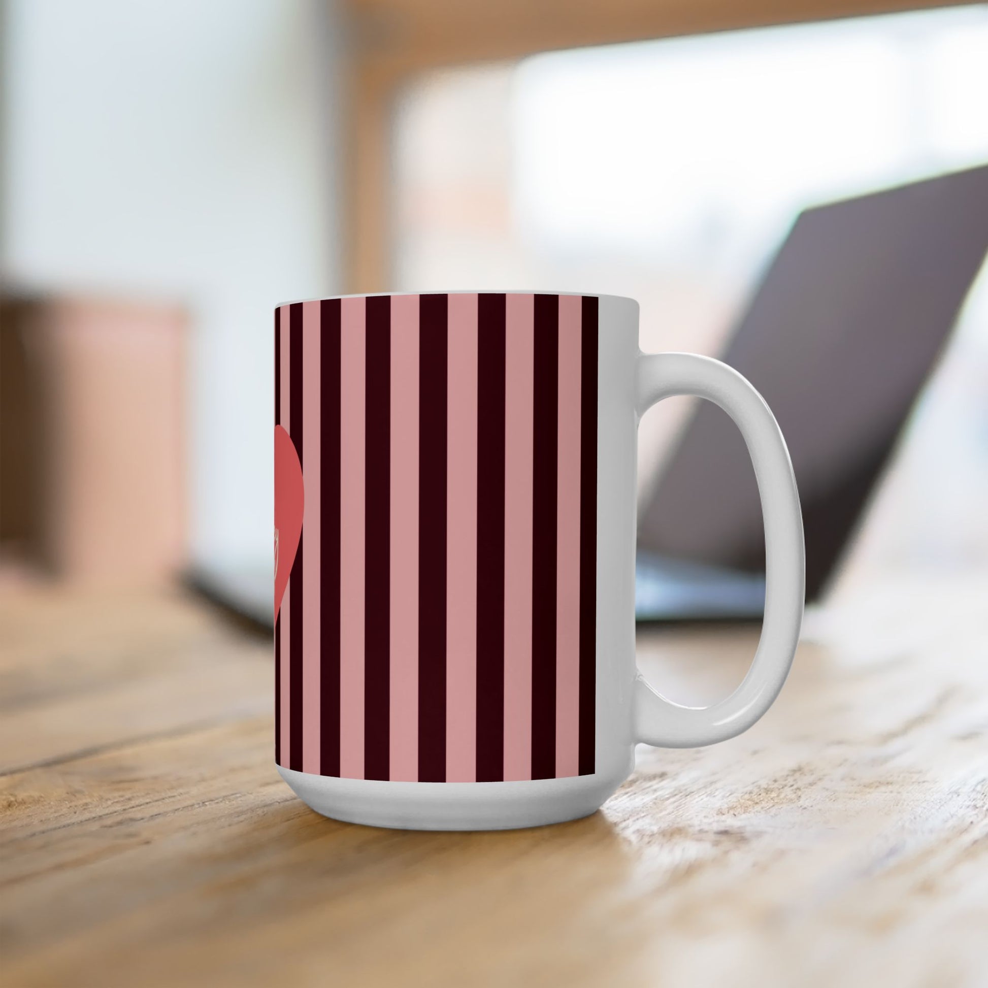 A white ceramic mug with vertical dark red and cream stripes, sitting on a wooden table with a blurred laptop in the background.