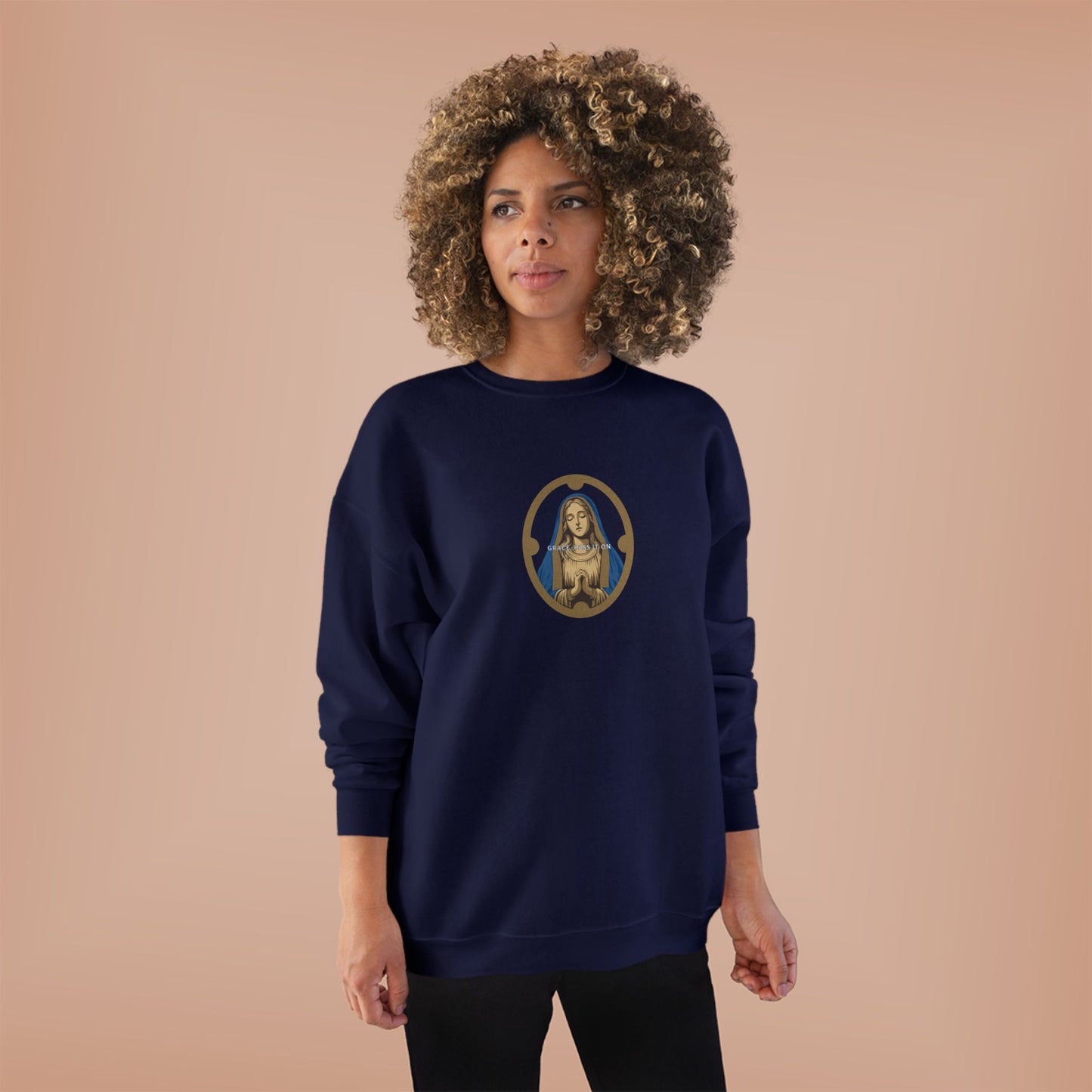 A woman with a voluminous curly afro stands against a solid peach studio background wearing a navy blue crewneck sweatshirt with a circular graphic of the Virgin Mary on the chest, paired with black pants.