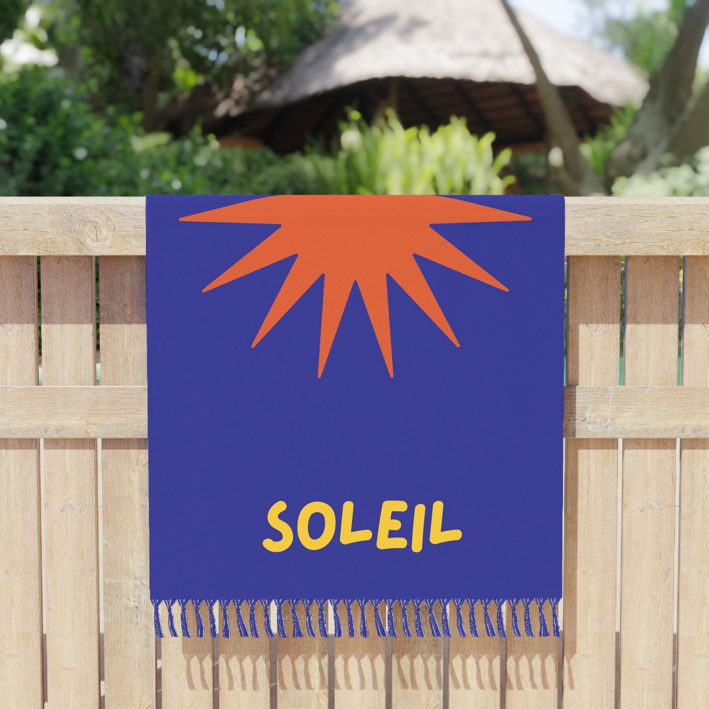 Towel with orange sun design and 'Soleil' text on a blue background, hanging on a wooden fence.