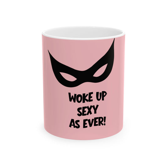 A pink ceramic mug with a black cat-eye mask graphic and the text 'WOKE UP SEXY AS EVER!' printed on the front, shown against a plain white background.