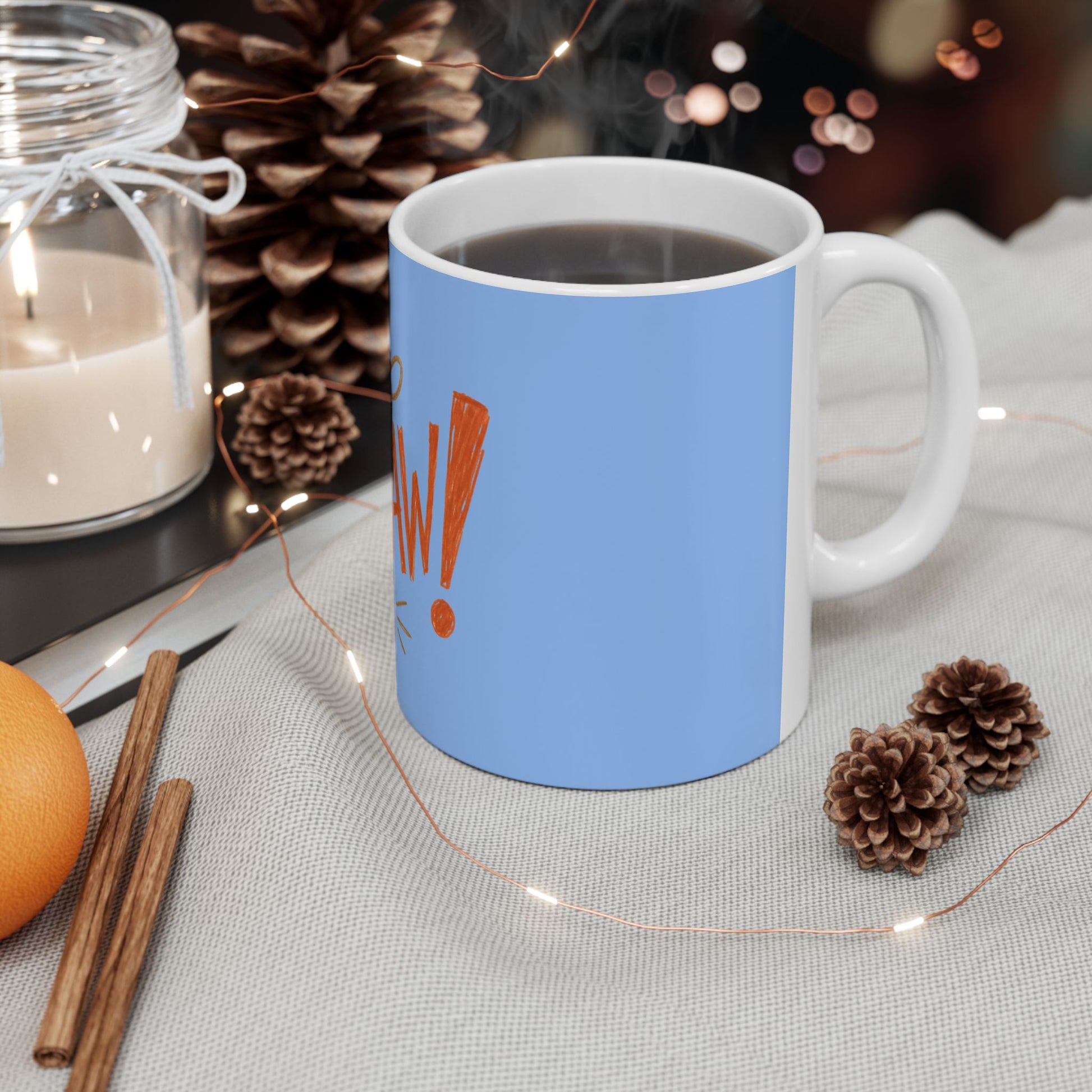 A light blue ceramic mug filled with a dark beverage sits on a light fabric surface, surrounded by holiday-style props including pinecones, a candle jar, cinnamon sticks, and fairy lights. The mug has a white side panel and handle, with an orange exclamation mark graphic on the front.