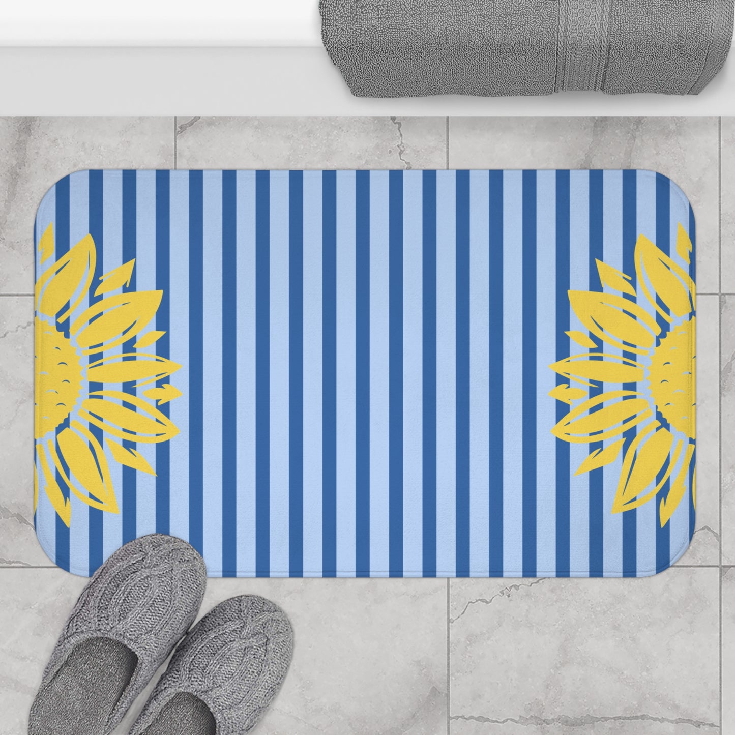 A rectangular bath mat with rounded corners, featuring a blue and light blue vertical stripe pattern with yellow sunflower designs on both sides, shown on a light marble floor with gray slippers and a folded gray towel nearby.