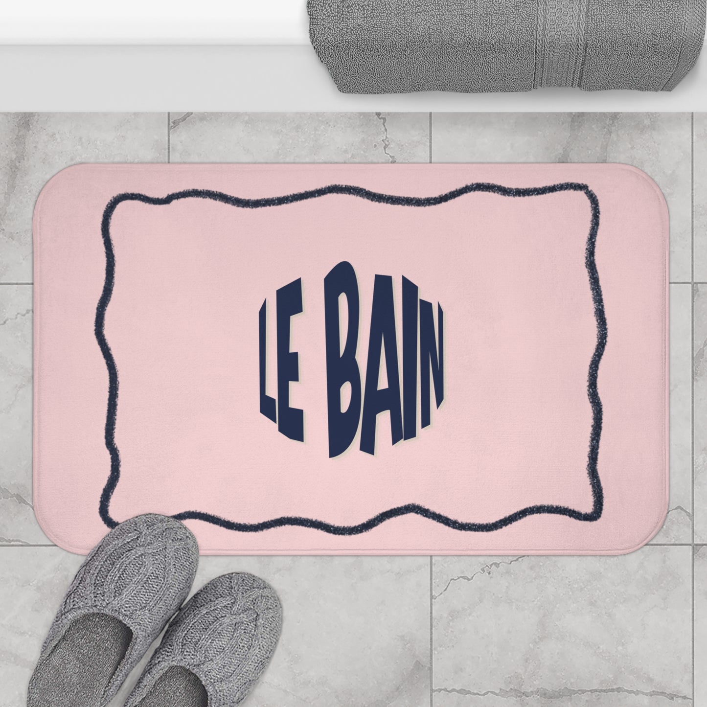 Pink bath mat with 'LE BAIN' text on a marble floor with slippers and a towel.