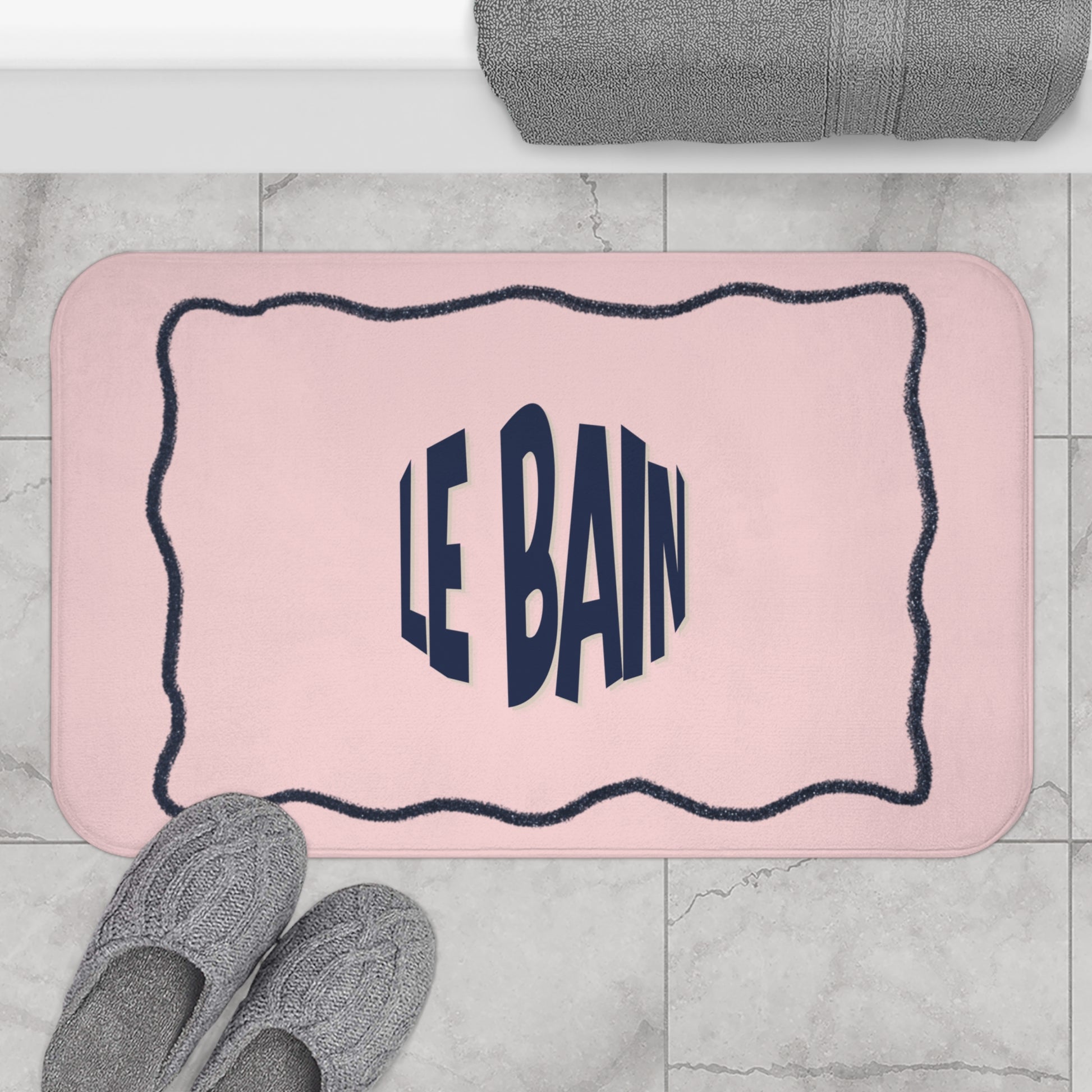 Pink bath mat with 'LE BAIN' text on a marble floor with slippers and a towel.
