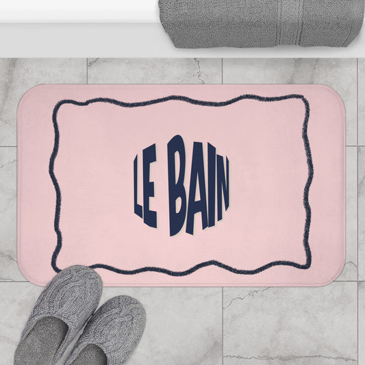 Pink bath mat with 'LE BAIN' text on a marble floor with slippers and a towel.