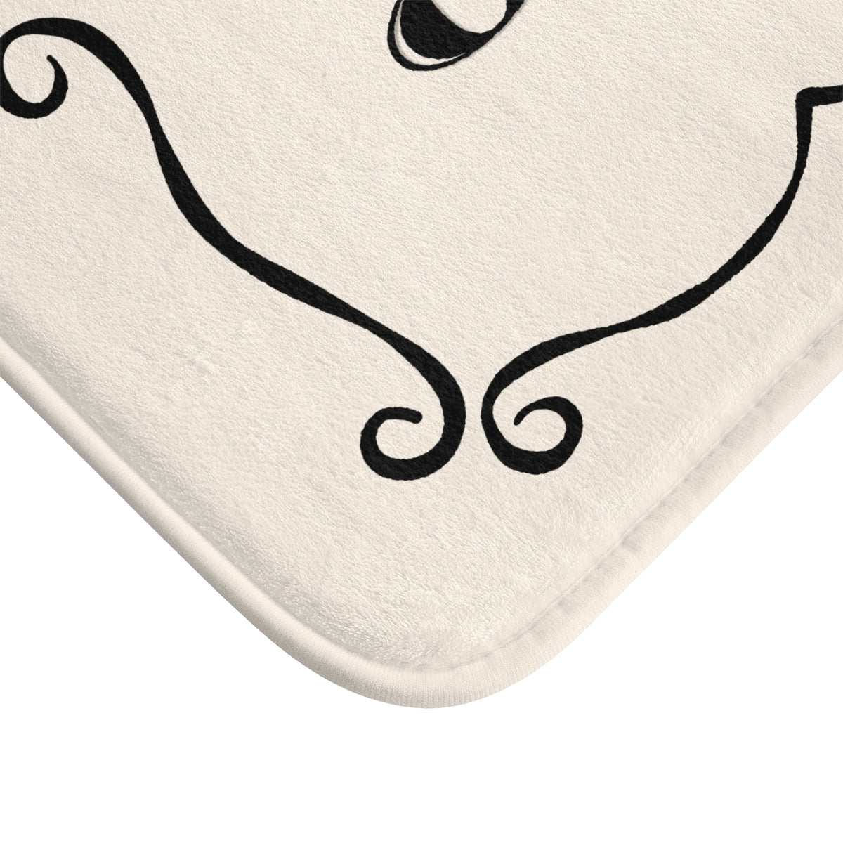 Close-up of a cream-colored bath mat with a black decorative swirl pattern, showing the corner and soft stitched edge against a white background.