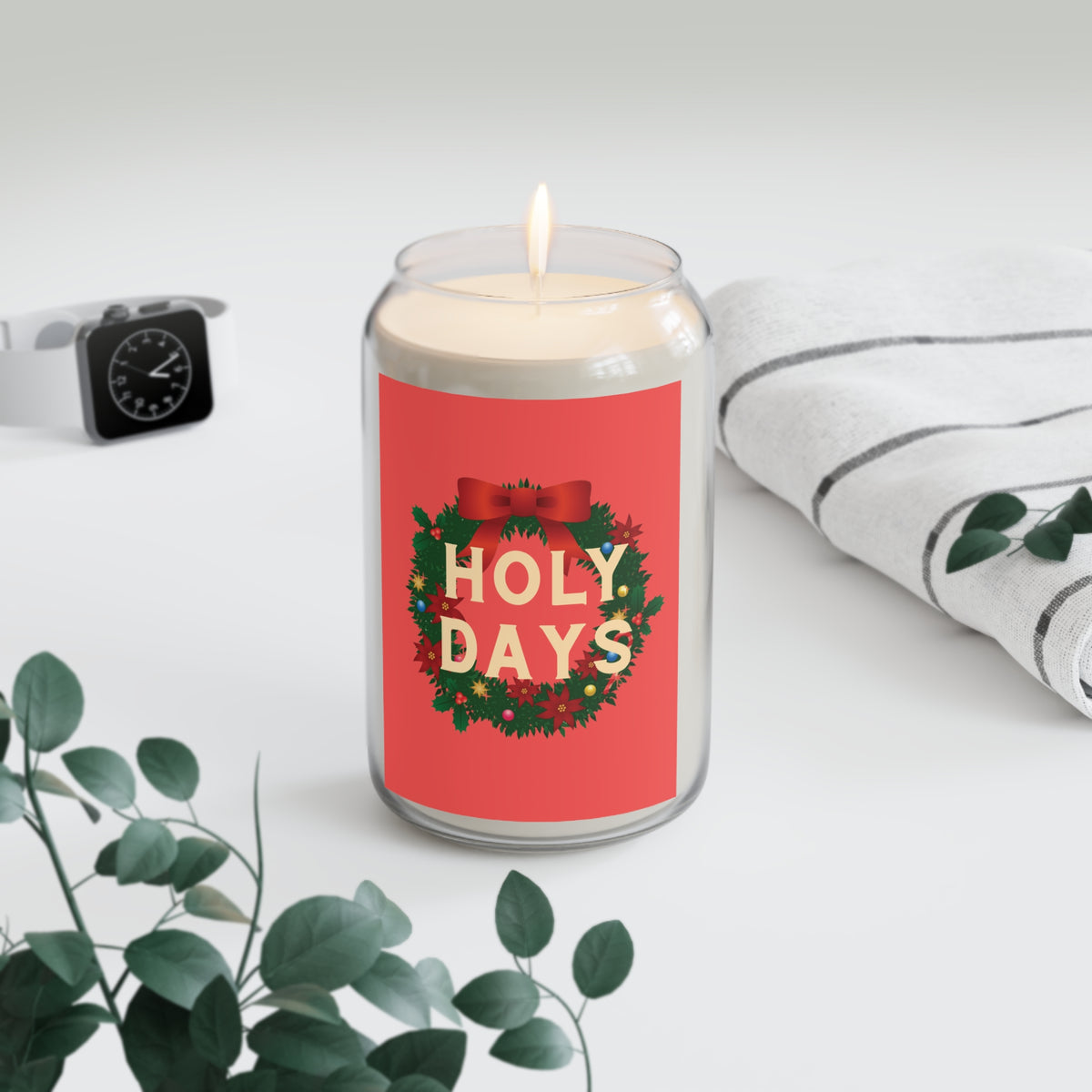 A lit glass jar candle with a red holiday label reading “HOLY DAYS” in gold letters, surrounded by a wreath design. The candle is styled on a light gray surface with a folded white striped towel, green leaves, and a black smartwatch in the background.