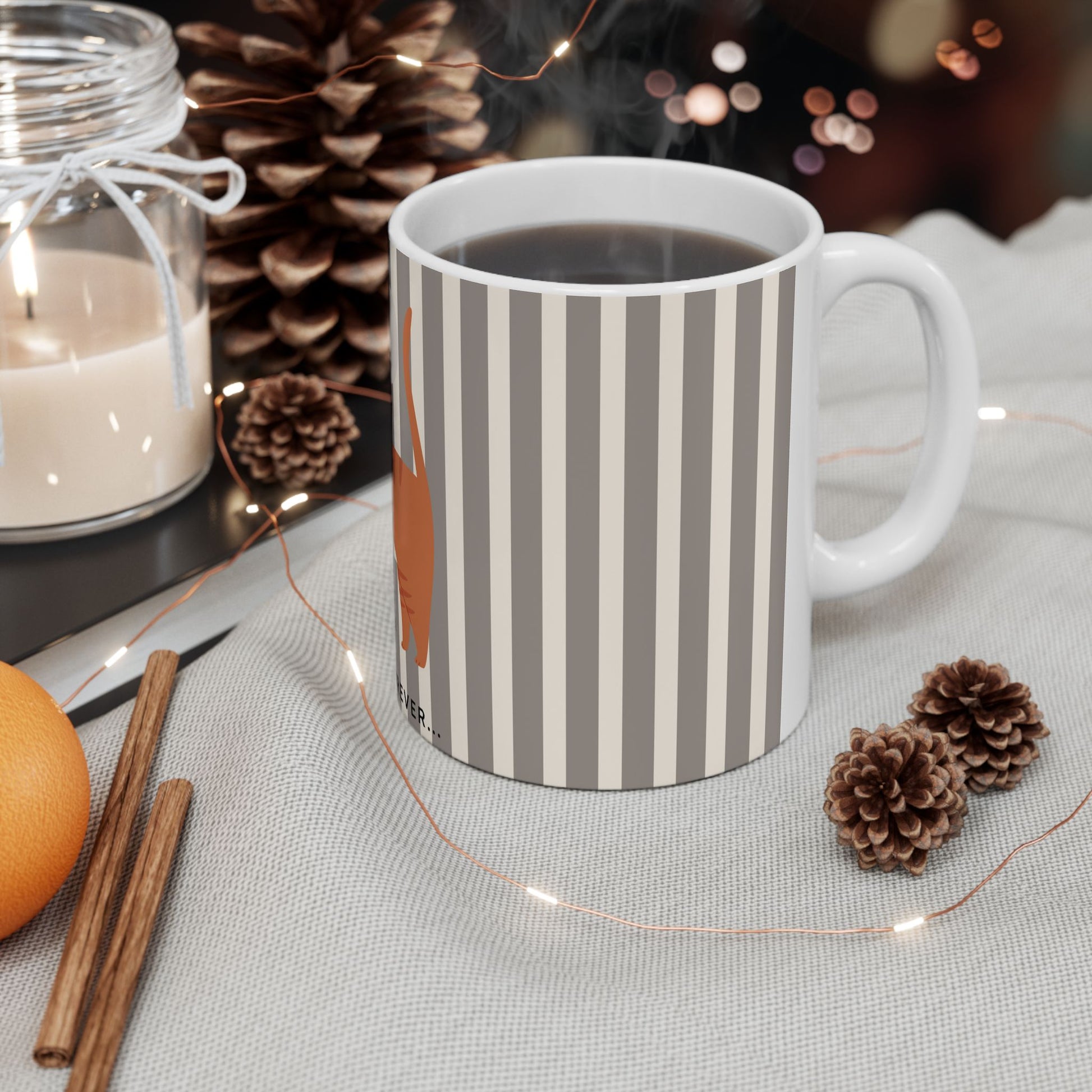 A white ceramic mug with vertical gray stripes and a stylized orange leaf graphic, filled with a dark beverage and placed on a light fabric surface with decorative string lights, pinecones, cinnamon sticks, an orange, and a candle in the background.