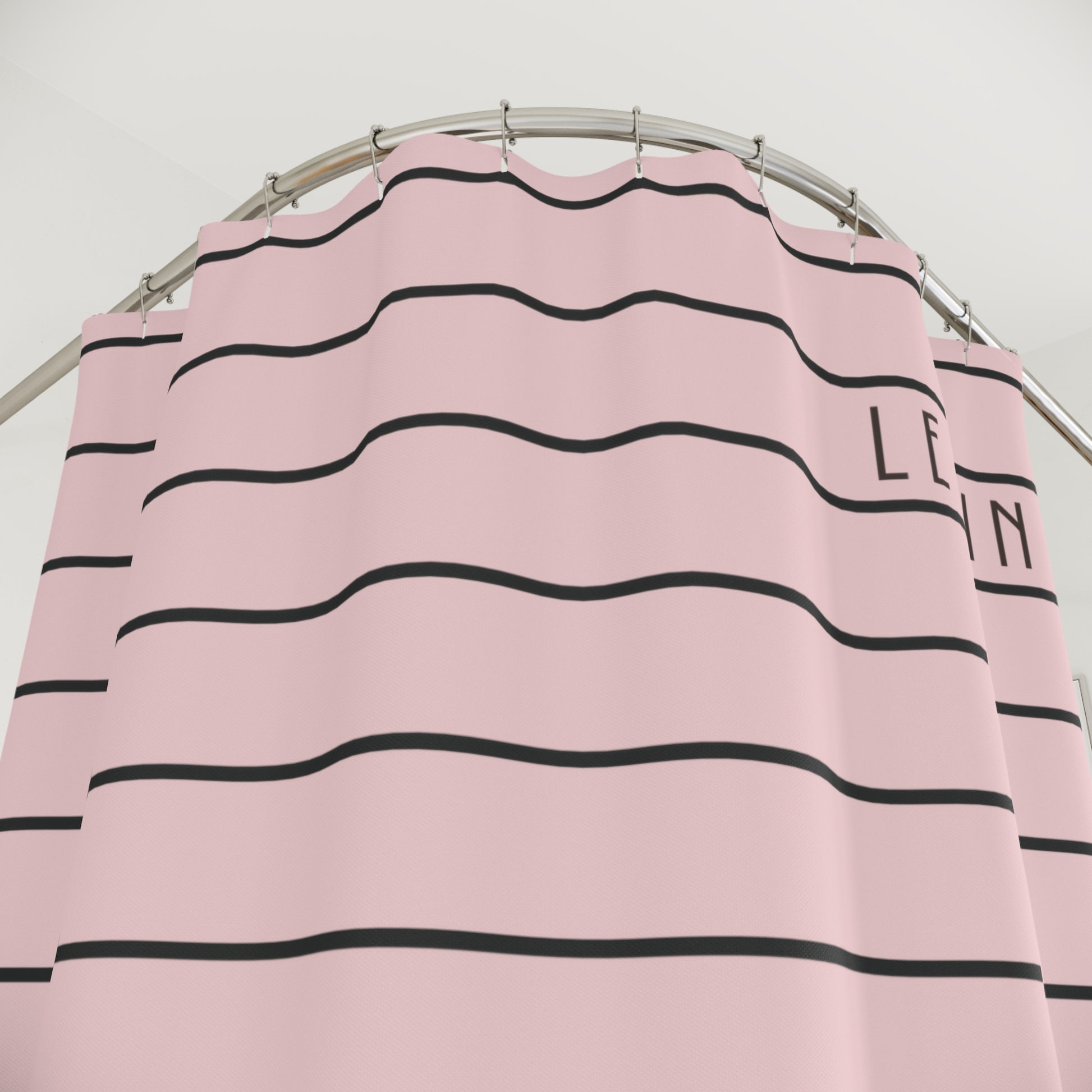 A pink shower curtain with horizontal black wavy stripes and partial black text on the right side, hanging from a curved silver metal rod against a white wall.