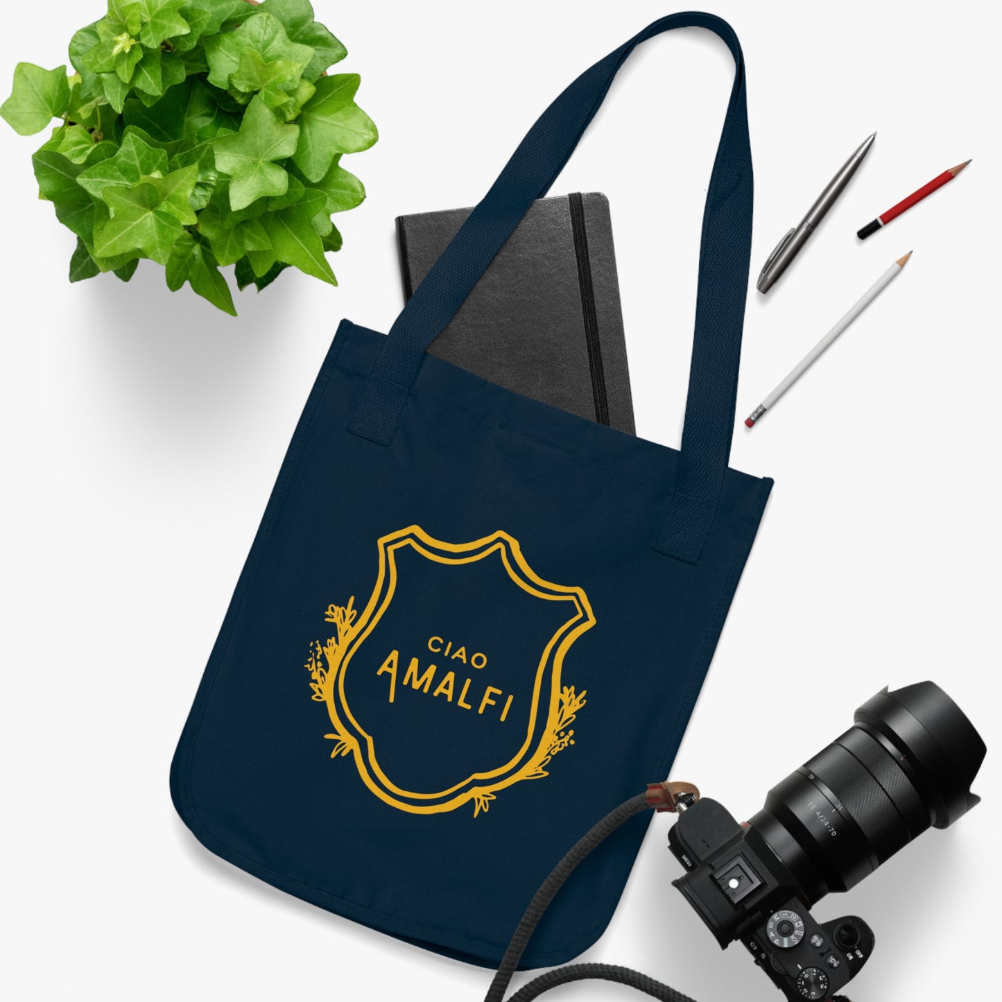 Black tote bag with a photo of a woman on a white surface with a plant, book, and camera.