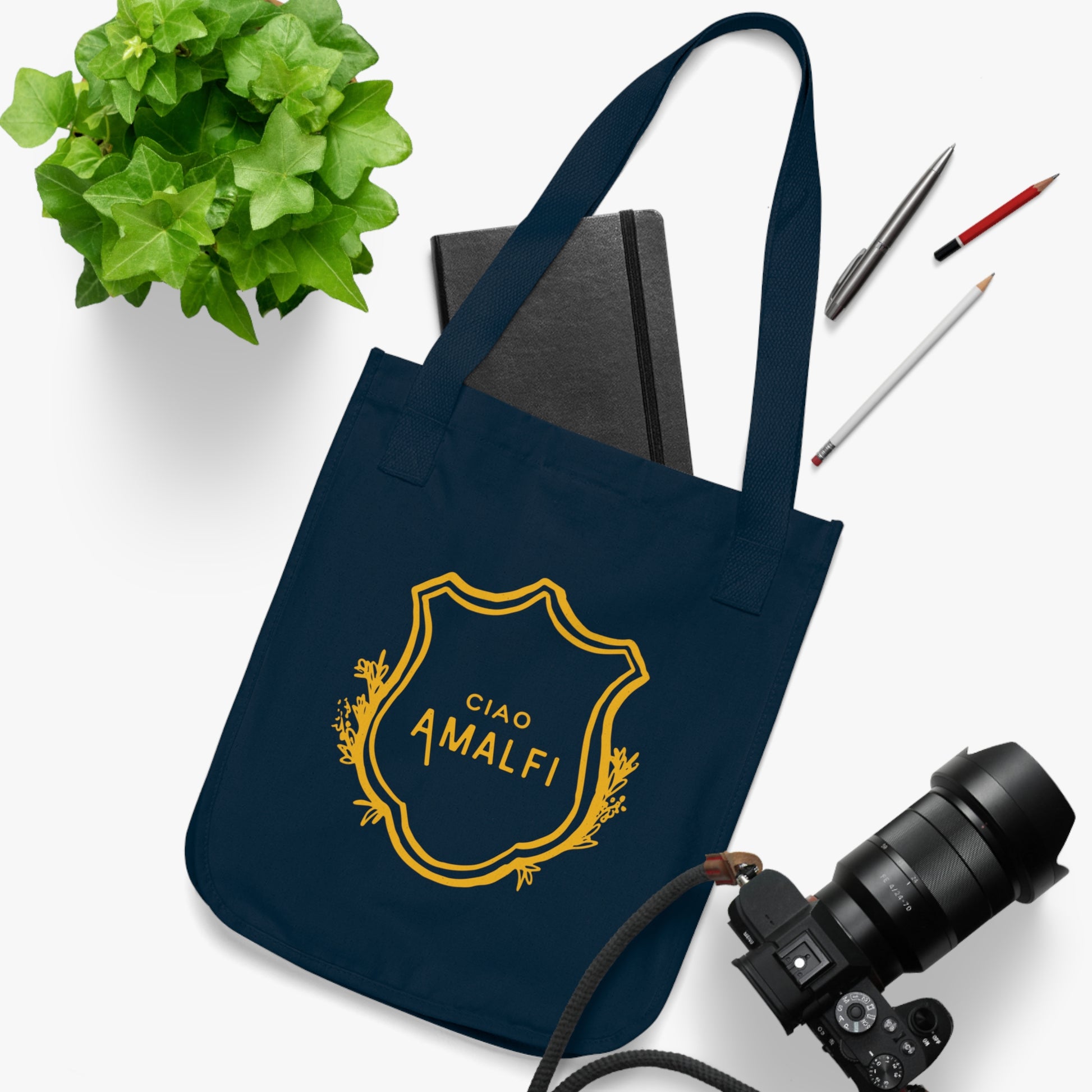 Black tote bag with a photo of a woman on a white surface with a plant, book, and camera.