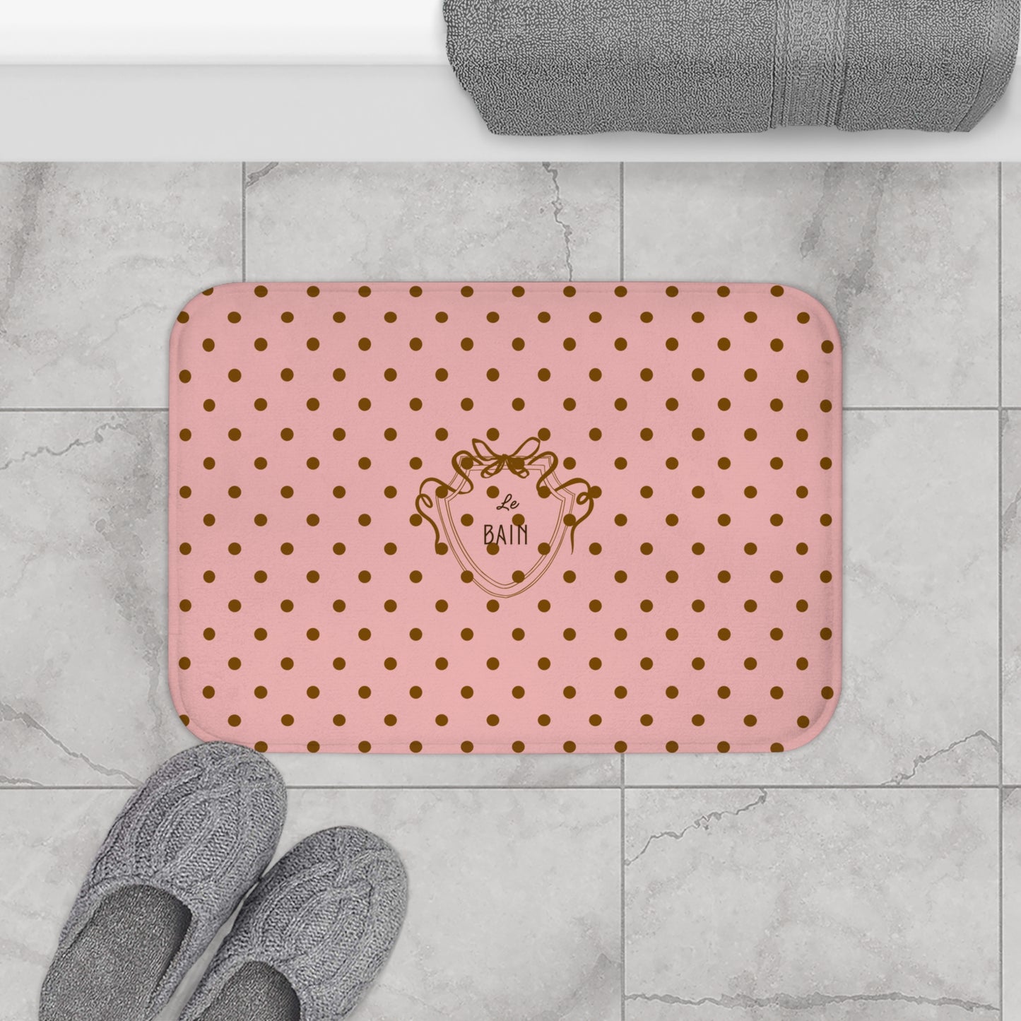 Pink bath mat with gold polka dots and a logo on a tiled bathroom floor.