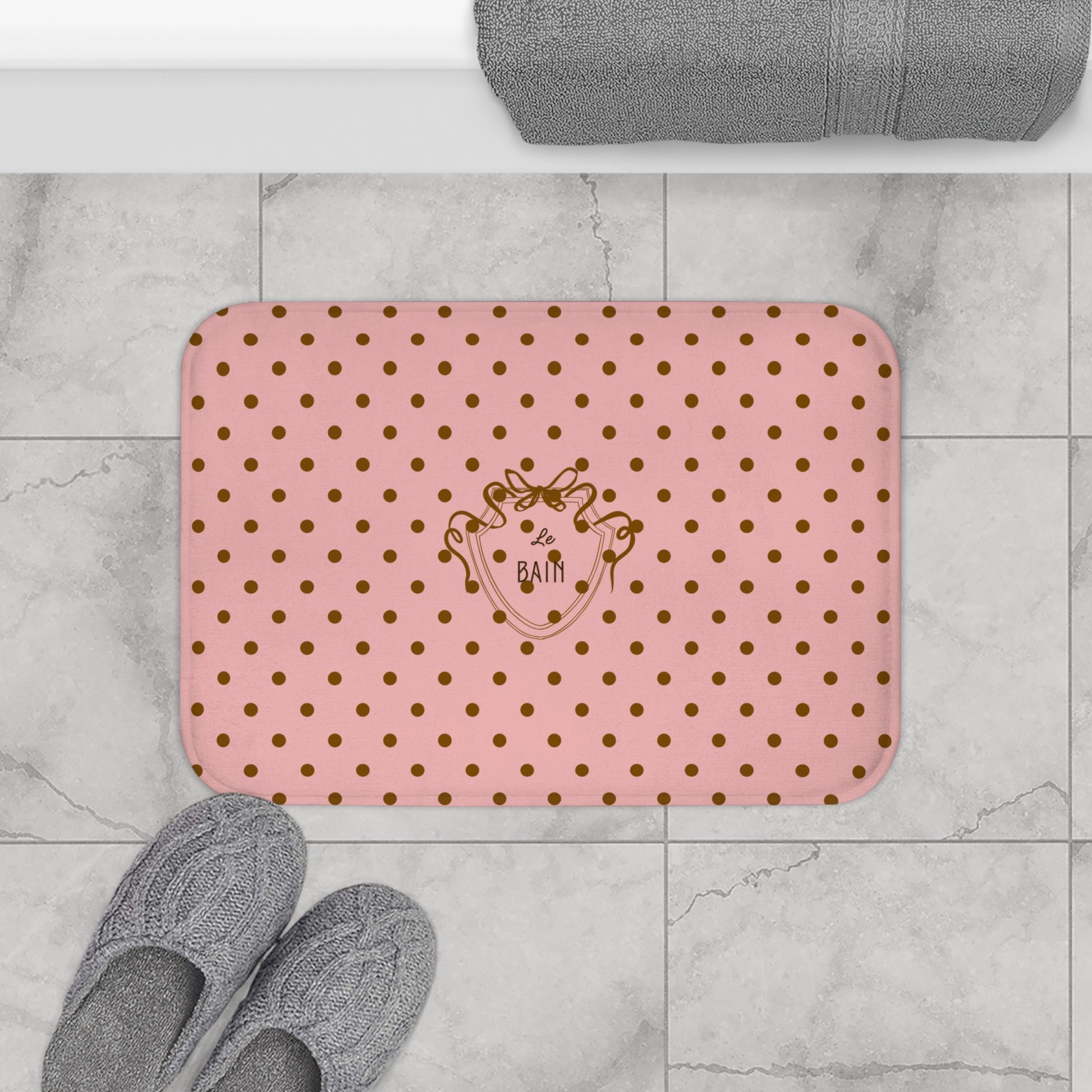 Pink bath mat with gold polka dots and a logo on a tiled bathroom floor.