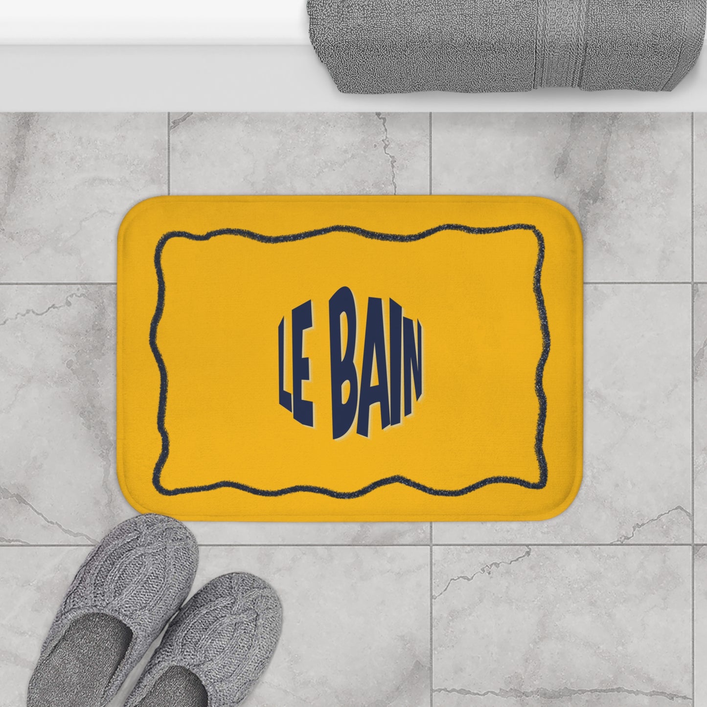Yellow bath mat with 'LE BAIN' text on a marble floor with gray slippers and a towel.