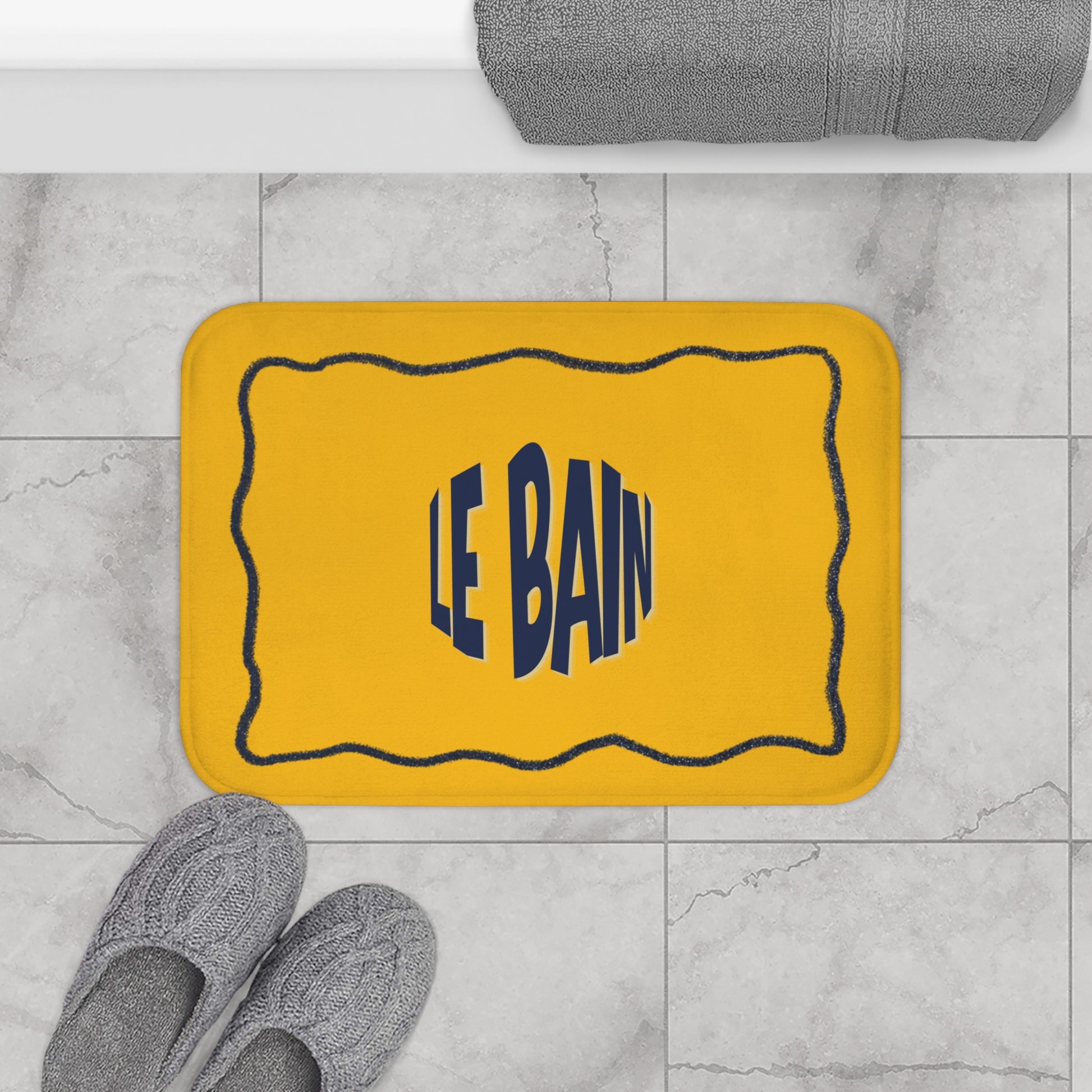 Yellow bath mat with 'LE BAIN' text on a marble floor with gray slippers and a towel.