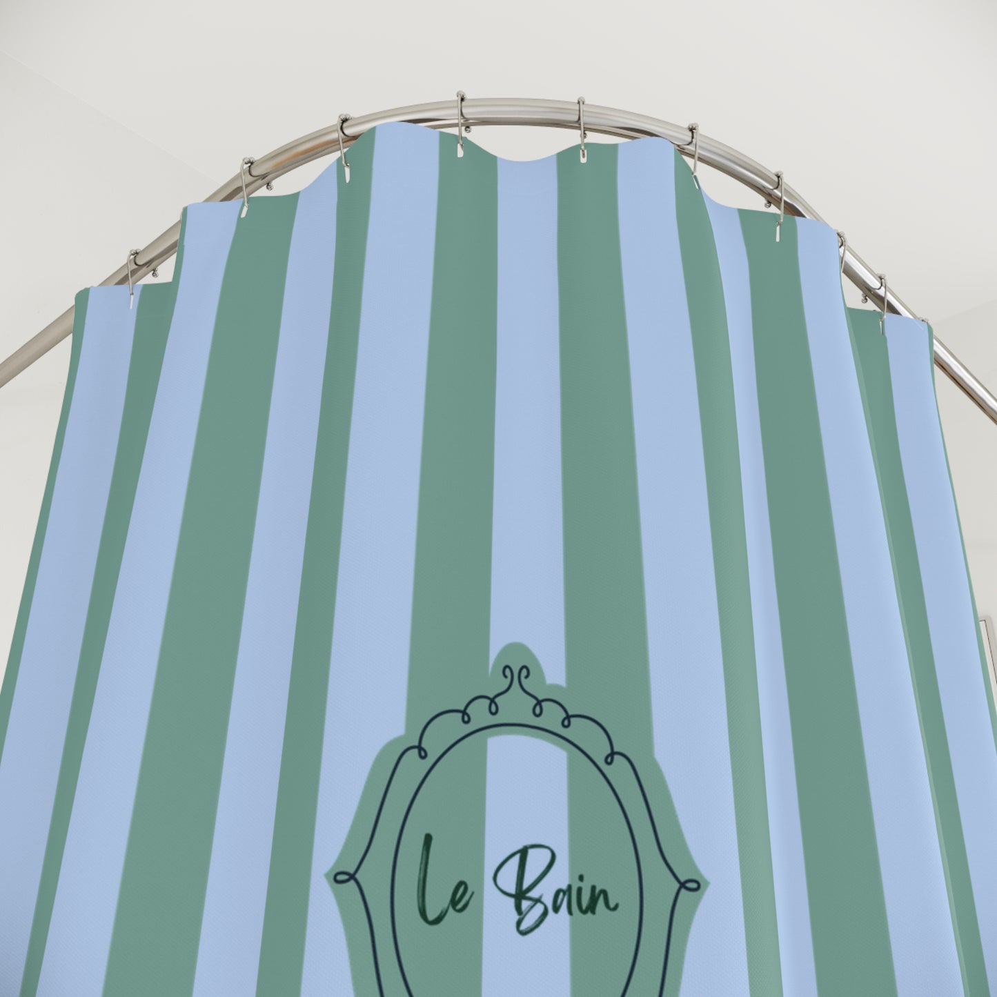 A striped shower curtain with alternating light blue and sage green vertical stripes, featuring a black ornate frame graphic with the text 'Le Bain' near the bottom center. The curtain is hanging from a curved metal rod against a plain off-white wall.