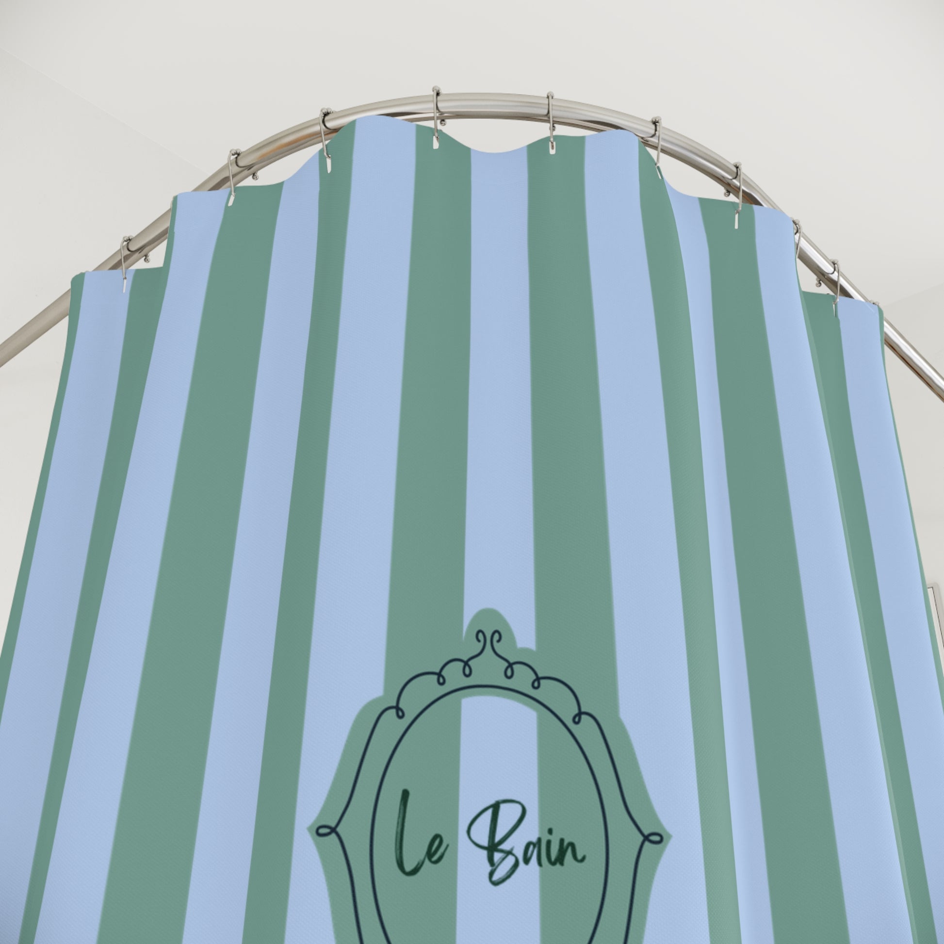 A striped shower curtain with alternating light blue and sage green vertical stripes, featuring a black ornate frame graphic with the text 'Le Bain' near the bottom center. The curtain is hanging from a curved metal rod against a plain off-white wall.