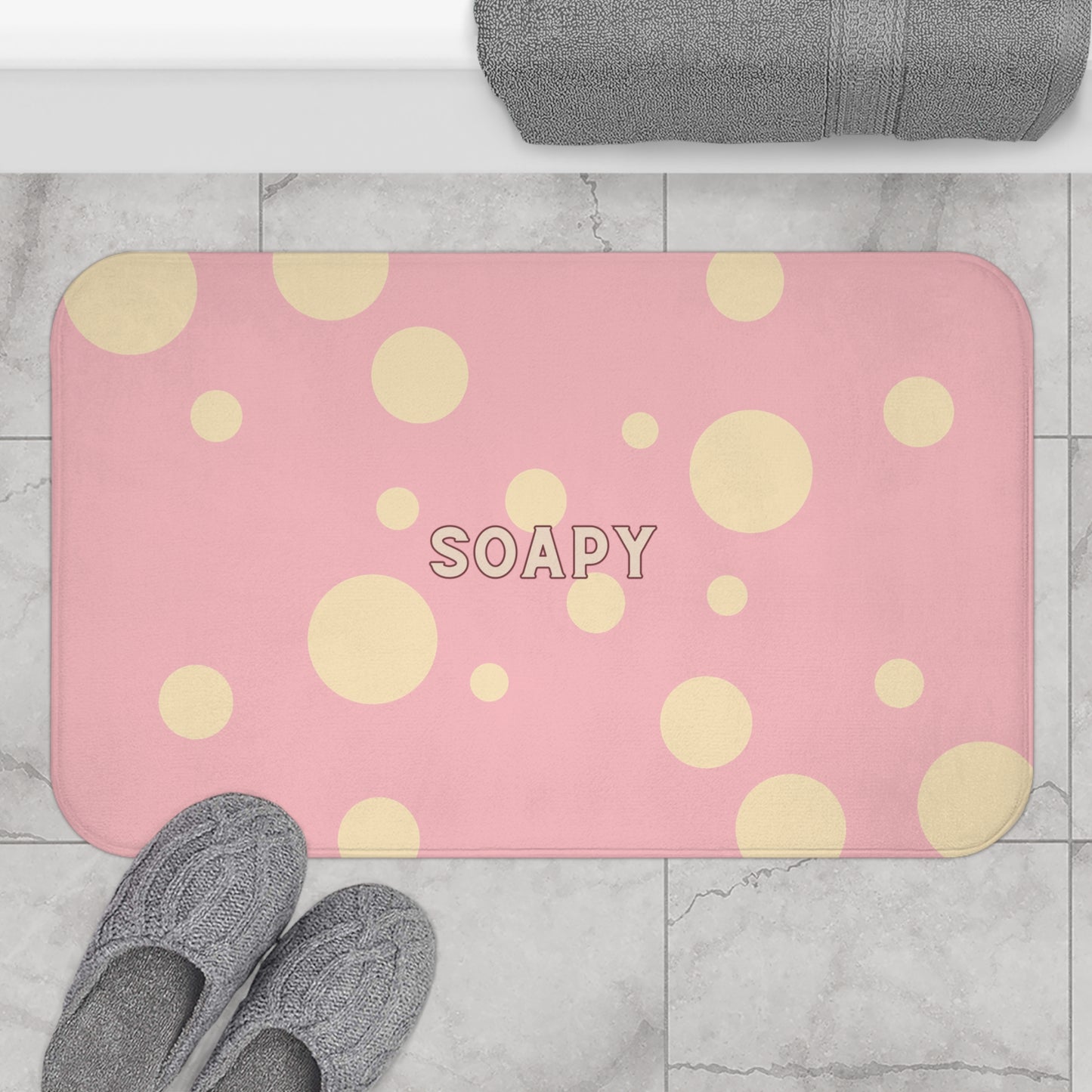 A pink rectangular bath mat with a yellow polka dot pattern and the word 'SOAPY' in the center, shown on a light marble tile floor with gray towels and slippers nearby.