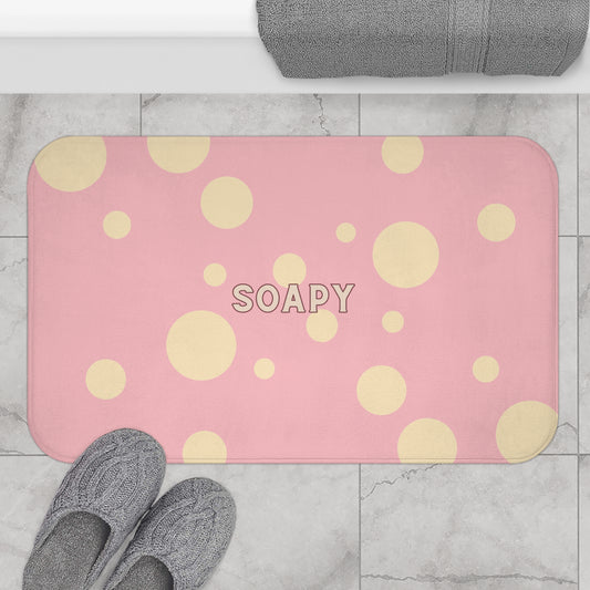A pink rectangular bath mat with a yellow polka dot pattern and the word 'SOAPY' in the center, shown on a light marble tile floor with gray towels and slippers nearby.