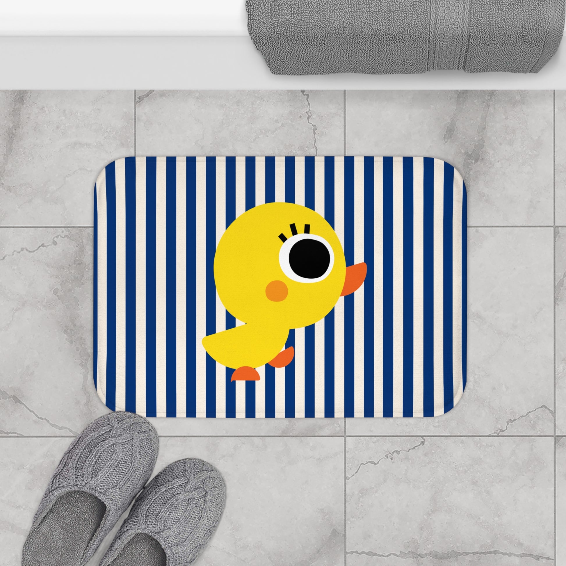 Bath mat with a yellow duck design on a blue and white striped background, placed on a tiled floor.