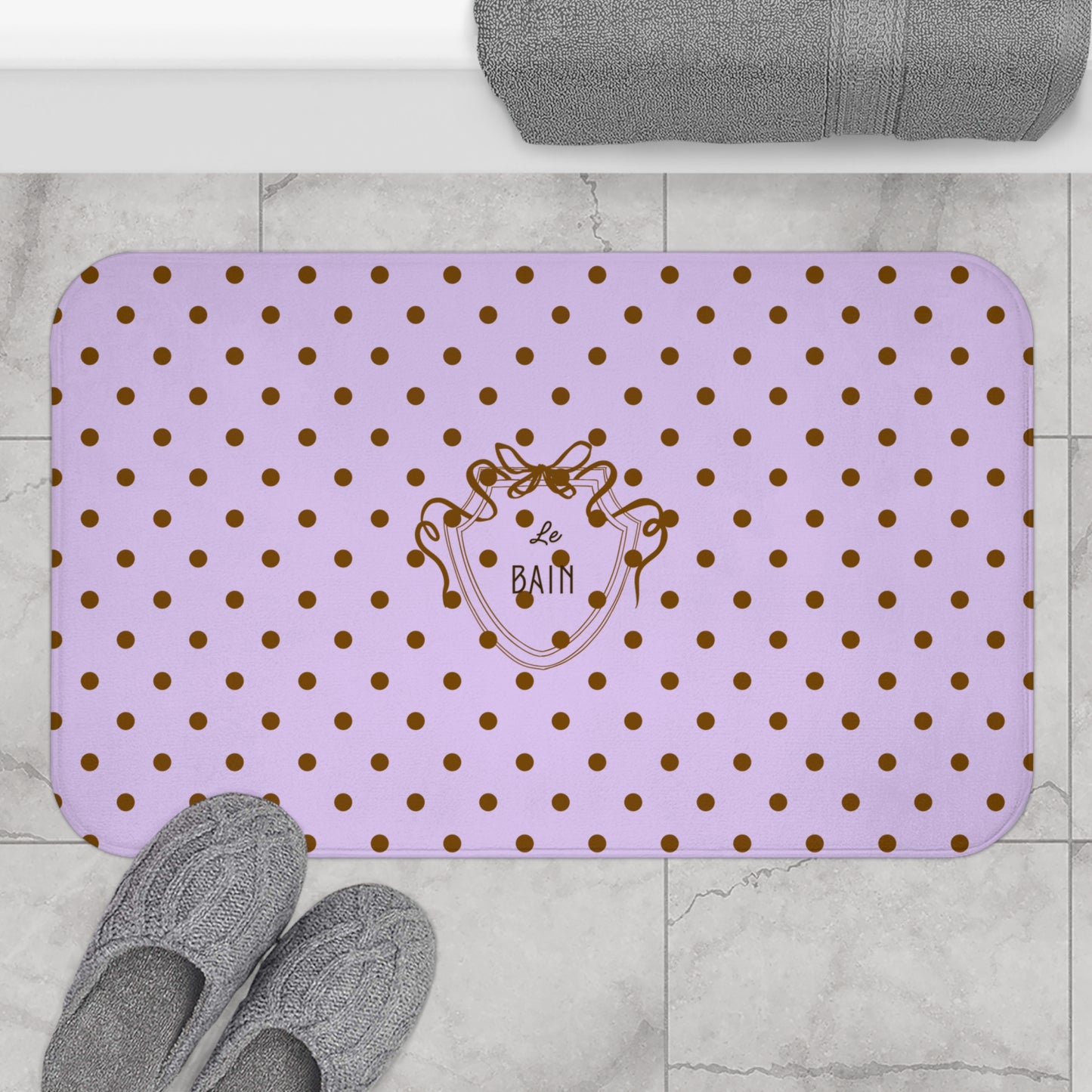 Purple bath mat with gold polka dots and a logo on a bathroom floor.