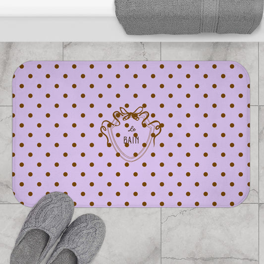 Purple bath mat with gold polka dots and a logo on a bathroom floor.