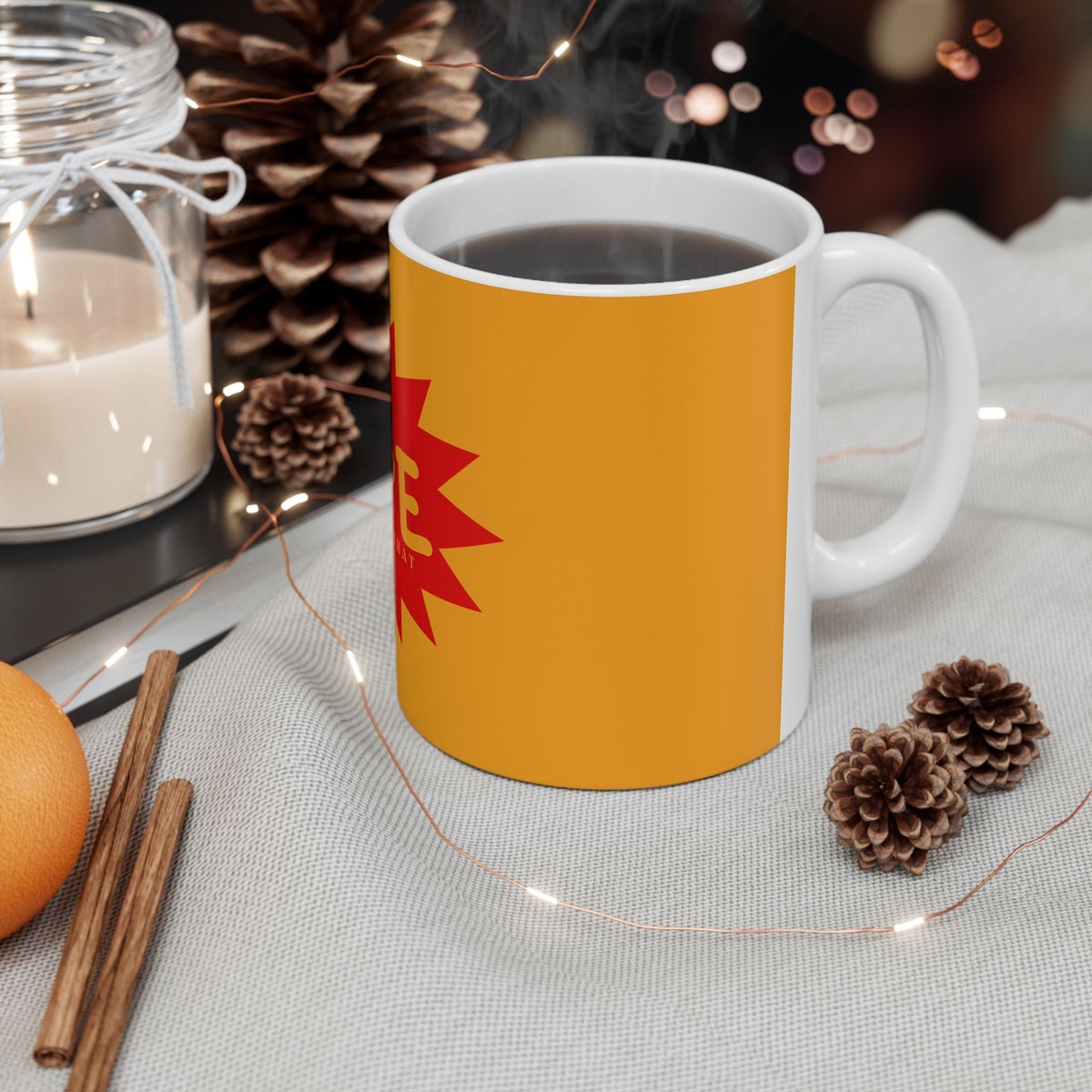 A white ceramic mug filled with a dark hot beverage, featuring a yellow-orange wraparound design with a red geometric graphic on the side. The mug is styled on a light gray fabric surface with pinecones, fairy lights, cinnamon sticks, an orange, and a glass jar candle in the background.