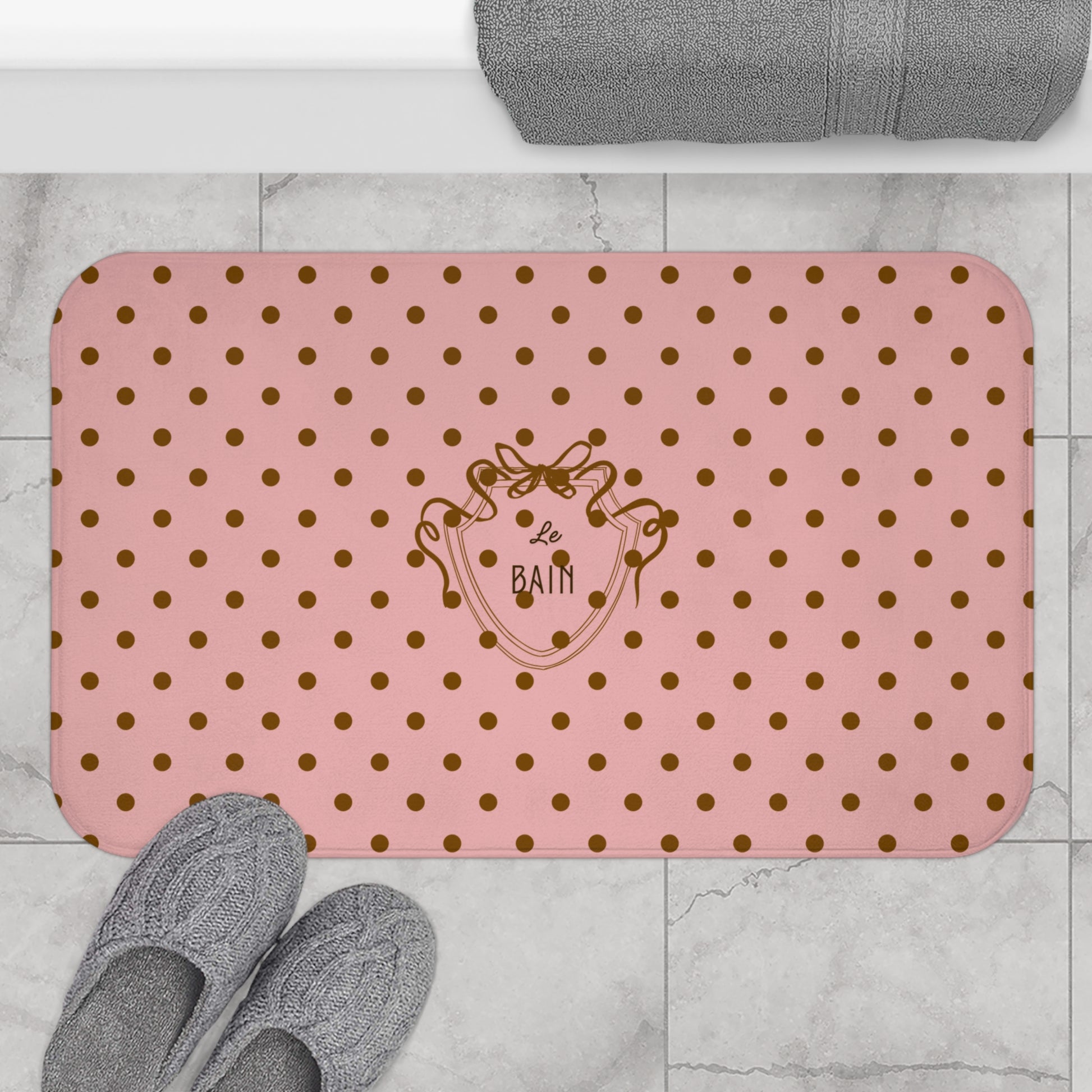 Pink bath mat with gold polka dots and a logo on a bathroom floor.