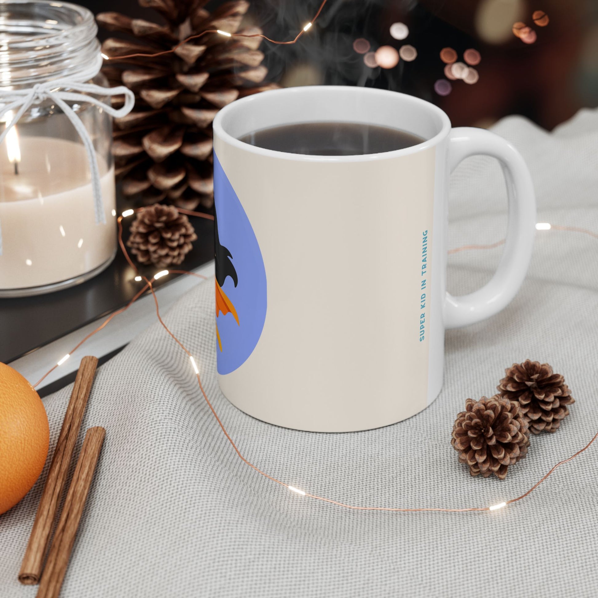 A white ceramic coffee mug filled with black coffee sits on a light fabric surface, surrounded by pinecones, a lit candle in a glass jar, cinnamon sticks, an orange, and warm string lights. The mug has a blue circular graphic with a dark silhouette and orange shape, plus small vertical blue text reading 'COFFEE IS IN TRAINING'.