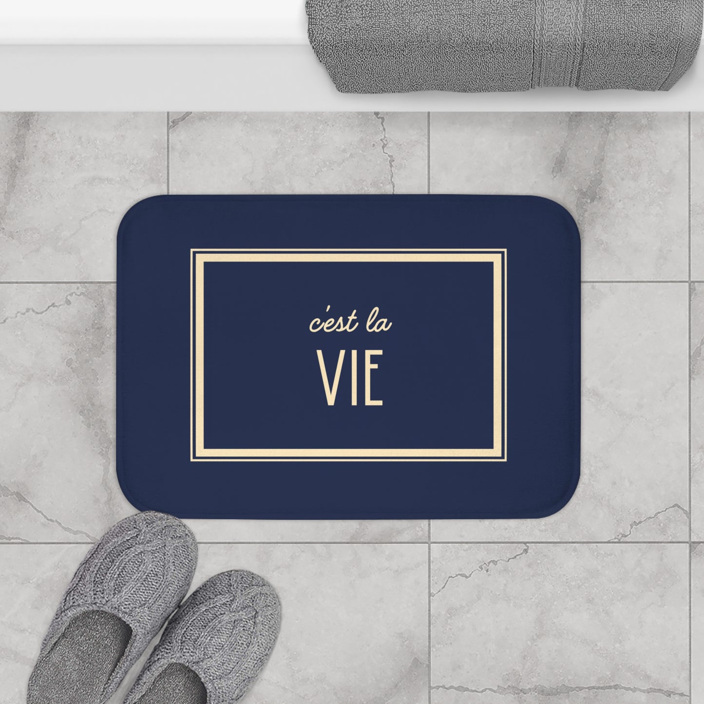 Navy blue doormat with 'c'est la Vie' text on a marble floor with slippers and a towel.