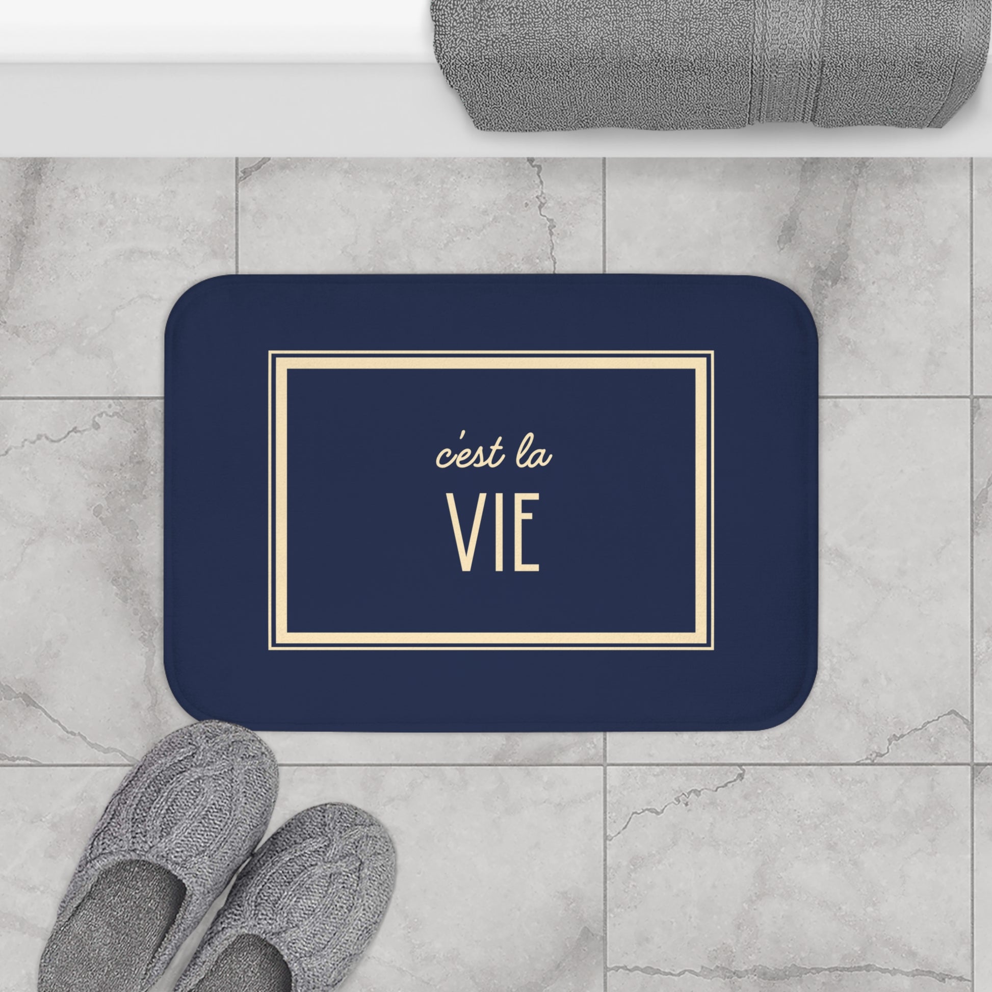 Navy blue doormat with 'c'est la Vie' text on a marble floor with slippers and a towel.