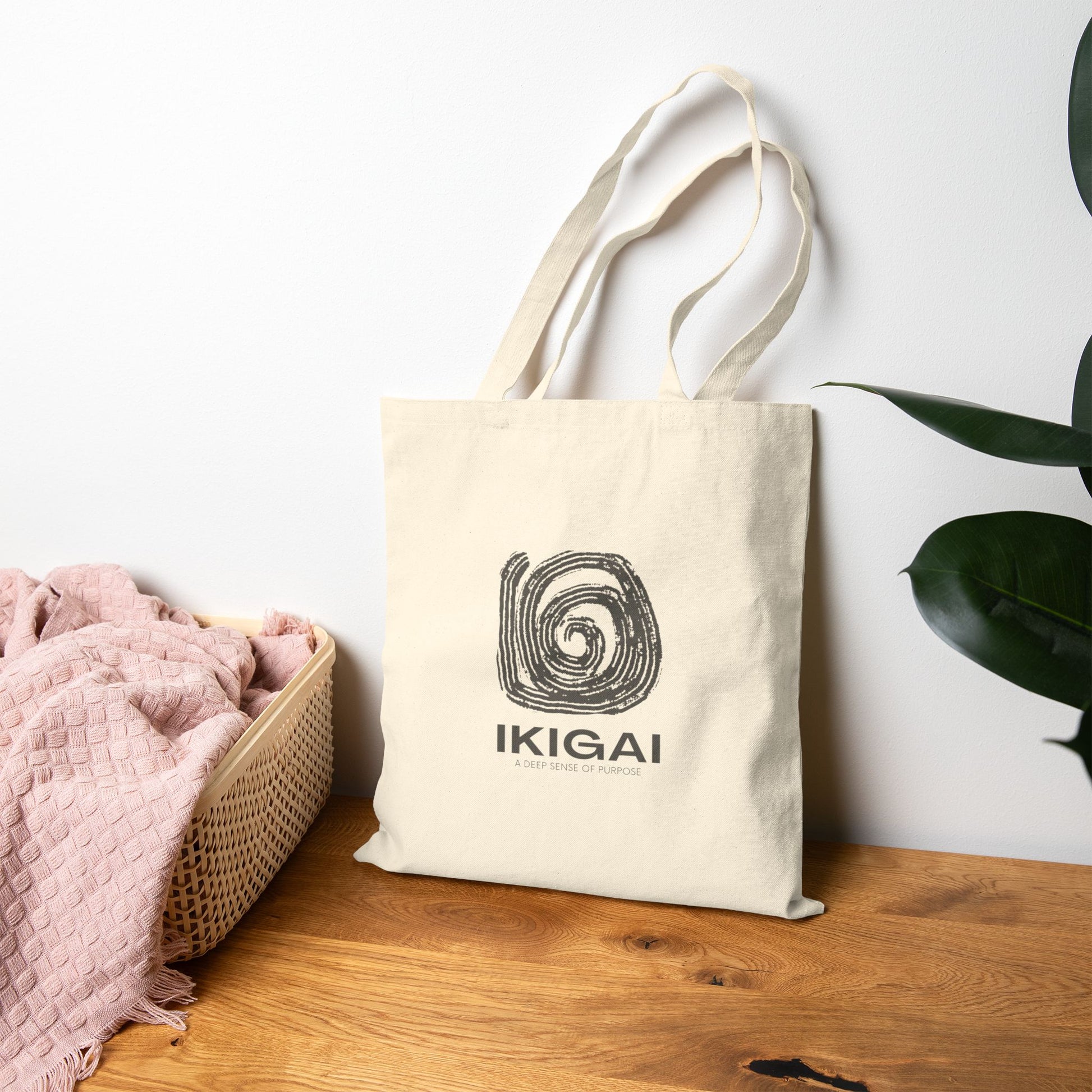 Beige tote bag with 'IKIGAI" design on a wooden surface with a white wall and plant in the background