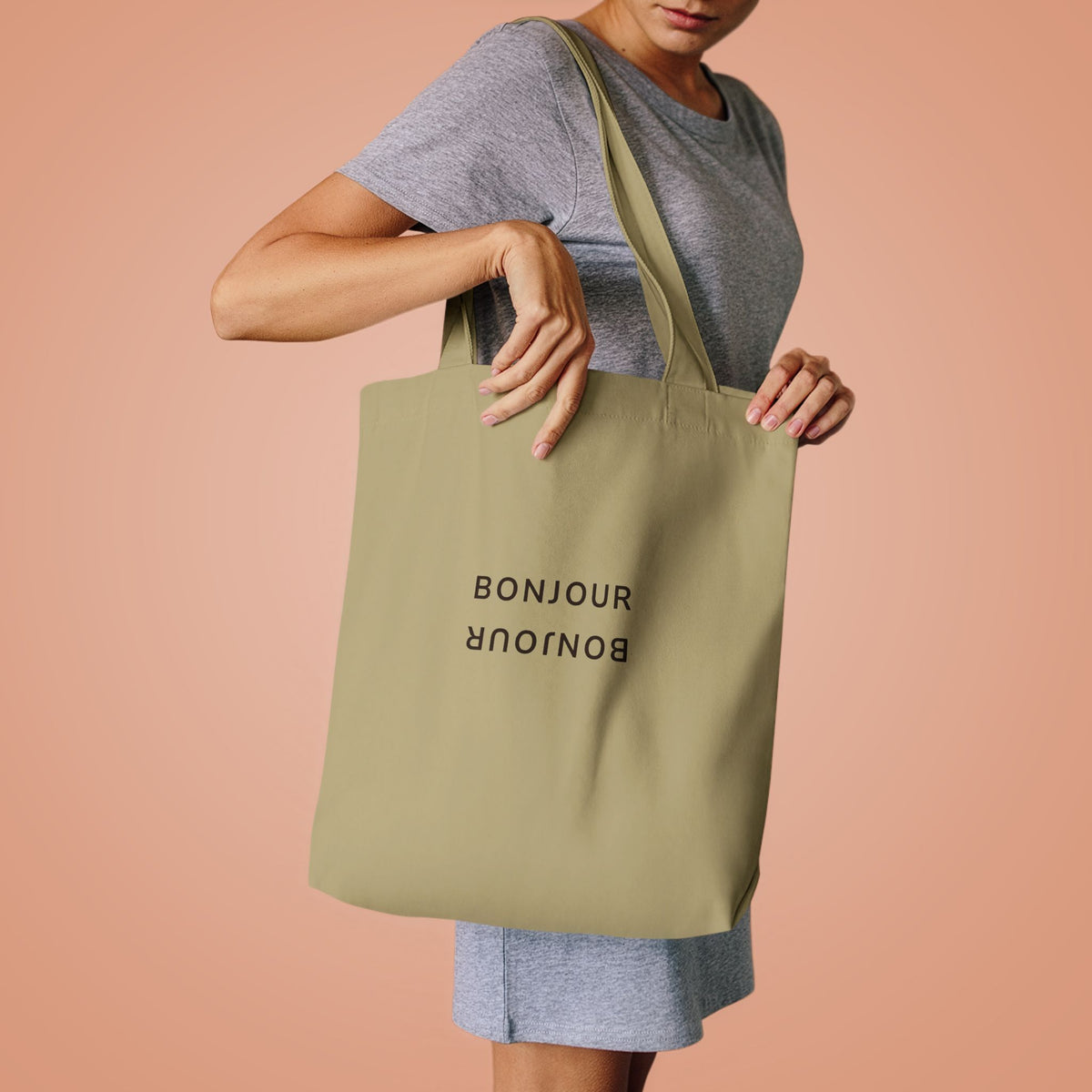 A woman wearing a gray t-shirt and shorts holds a sage green canvas tote bag with BONJOUR written on a peach-colored background.