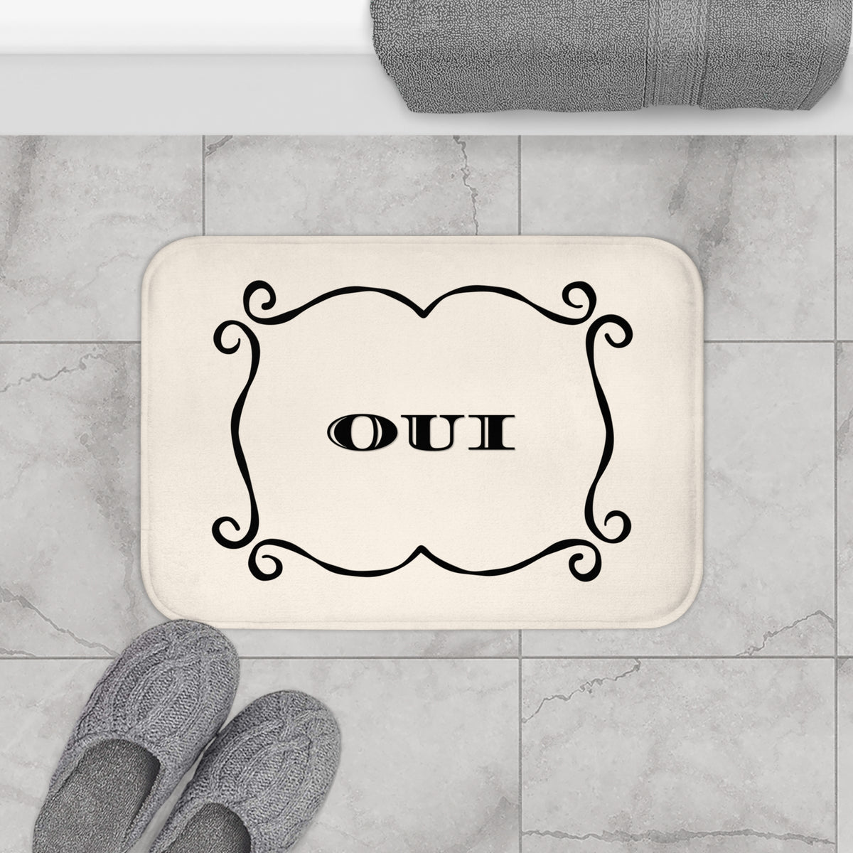 A rectangular cream bath mat with rounded corners sits on a light gray marble tile floor. The mat has a black ornamental border and the word 'OUI' in the center.