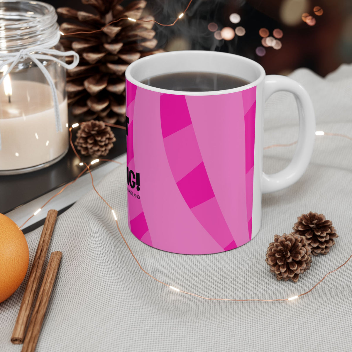 A white ceramic coffee mug with a bright pink geometric pattern, filled with black coffee and placed on a light gray fabric surface with holiday-themed props like pinecones, a candle, cinnamon sticks, and fairy lights.