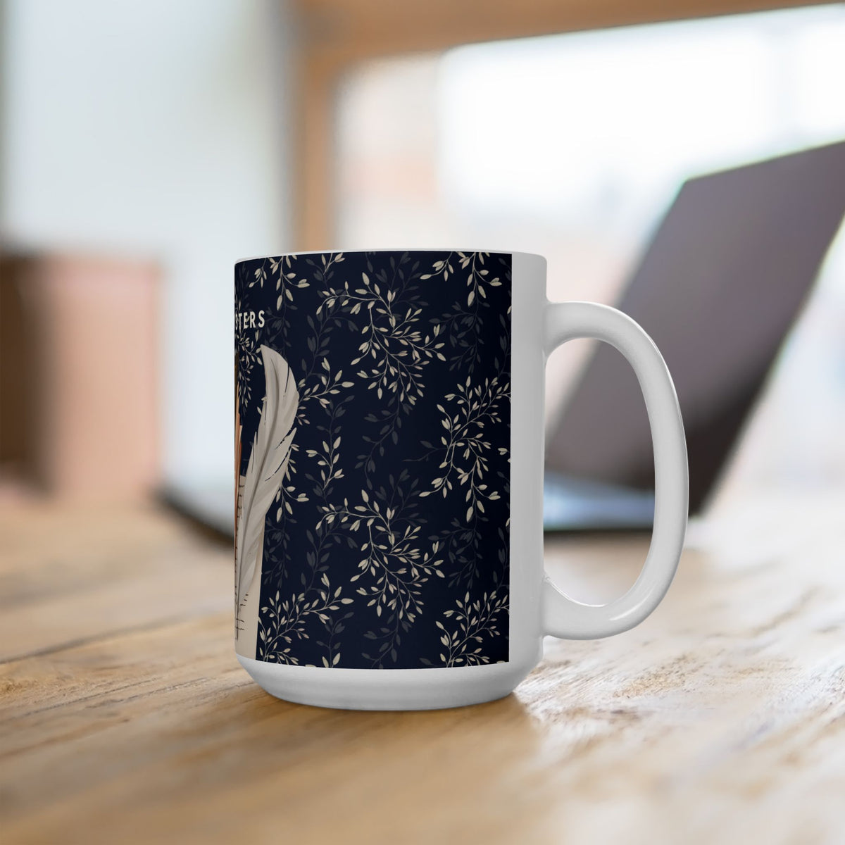 A white ceramic mug with a dark navy blue floral pattern and a white feather illustration, sitting on a wooden table with a blurred laptop in the background.