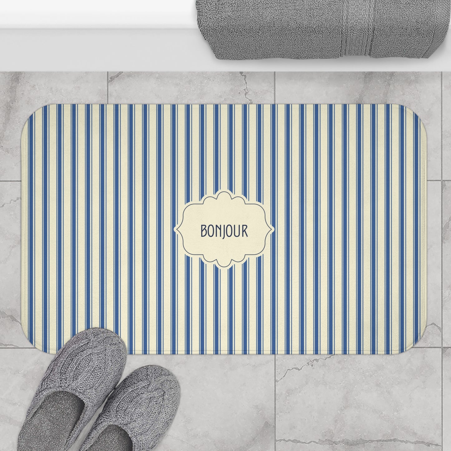 A rectangular bath mat with rounded corners, featuring vertical blue and cream stripes and a central cream label that reads 'BONJOUR'. The mat is styled in a bathroom setting on a light marble tile floor, with gray slippers at the bottom left and a folded gray towel at the top.