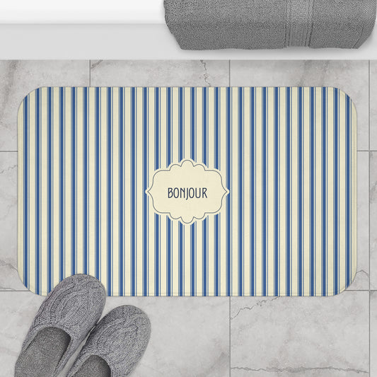 A rectangular bath mat with rounded corners, featuring vertical blue and cream stripes and a central cream label that reads 'BONJOUR'. The mat is styled in a bathroom setting on a light marble tile floor, with gray slippers at the bottom left and a folded gray towel at the top.
