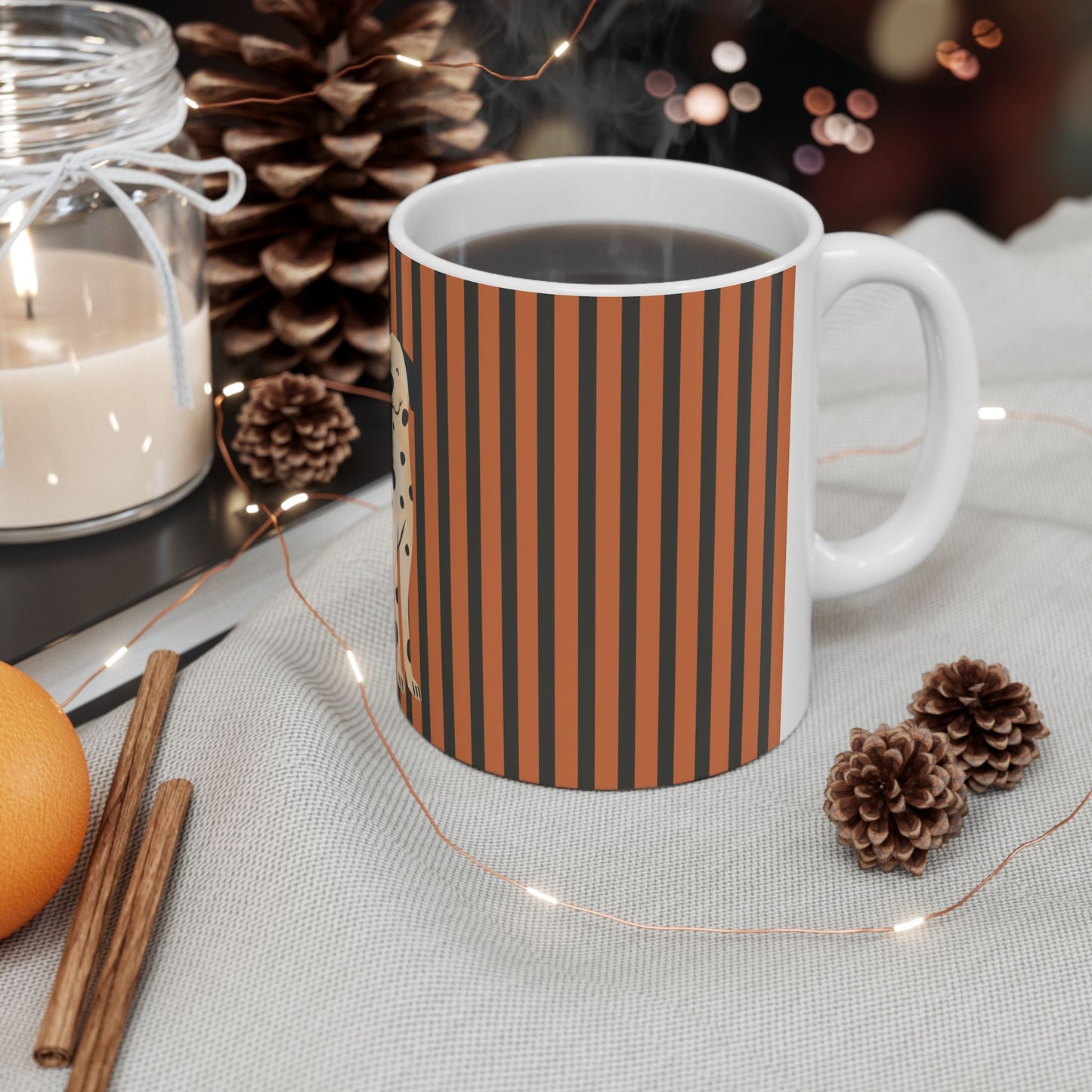 A white ceramic mug filled with a dark beverage, featuring vertical rust-orange and black stripes on the side, styled on a light fabric surface with pinecones, a candle jar, cinnamon sticks, and warm string lights.