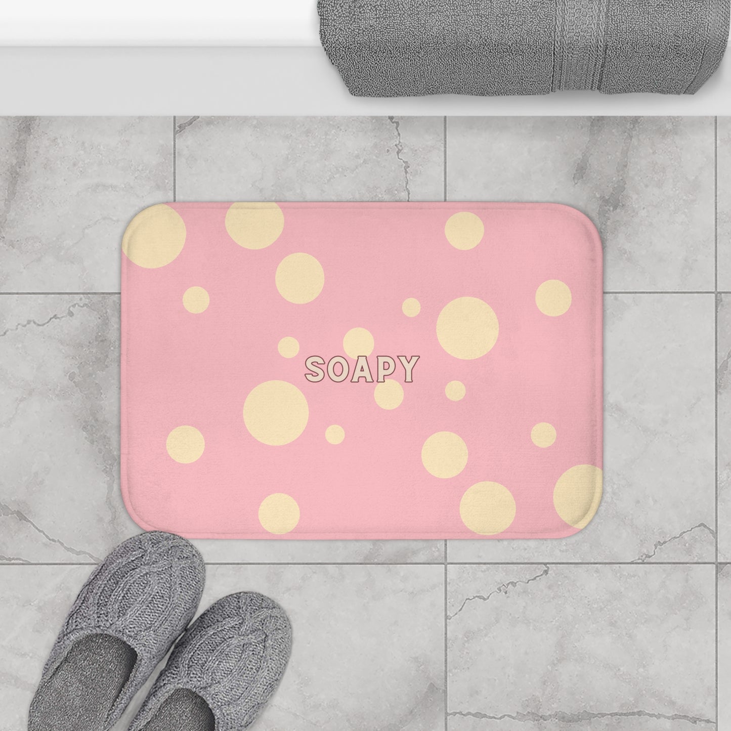 A pink rectangular bath mat with a yellow polka dot pattern and the word 'SOAPY' printed in the center, shown on a gray marble-look tiled floor with gray slippers and a folded gray towel nearby.