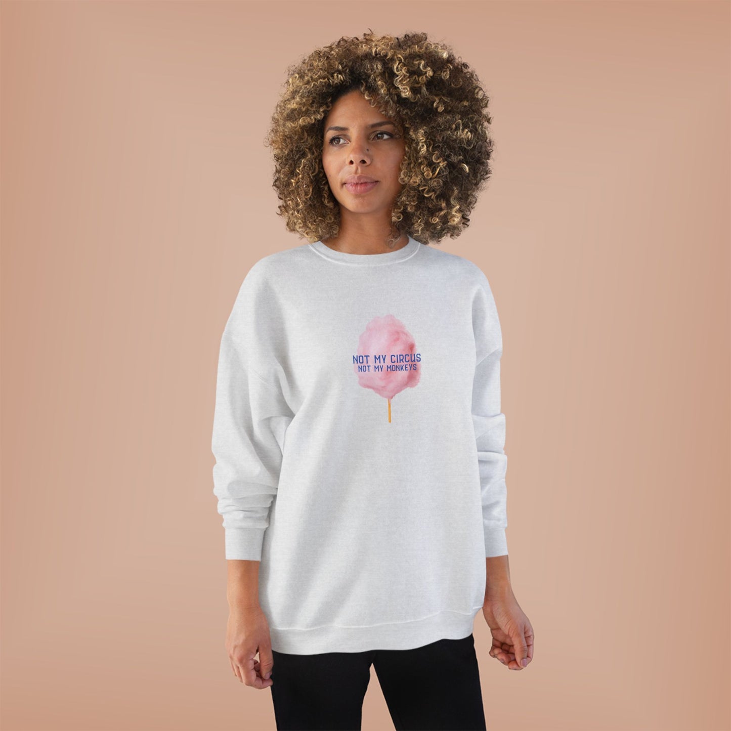 A woman with a large curly afro stands against a solid peach studio background wearing a light gray sweatshirt with a pink cotton candy graphic and the text 'NOT MY CIRCUS .
