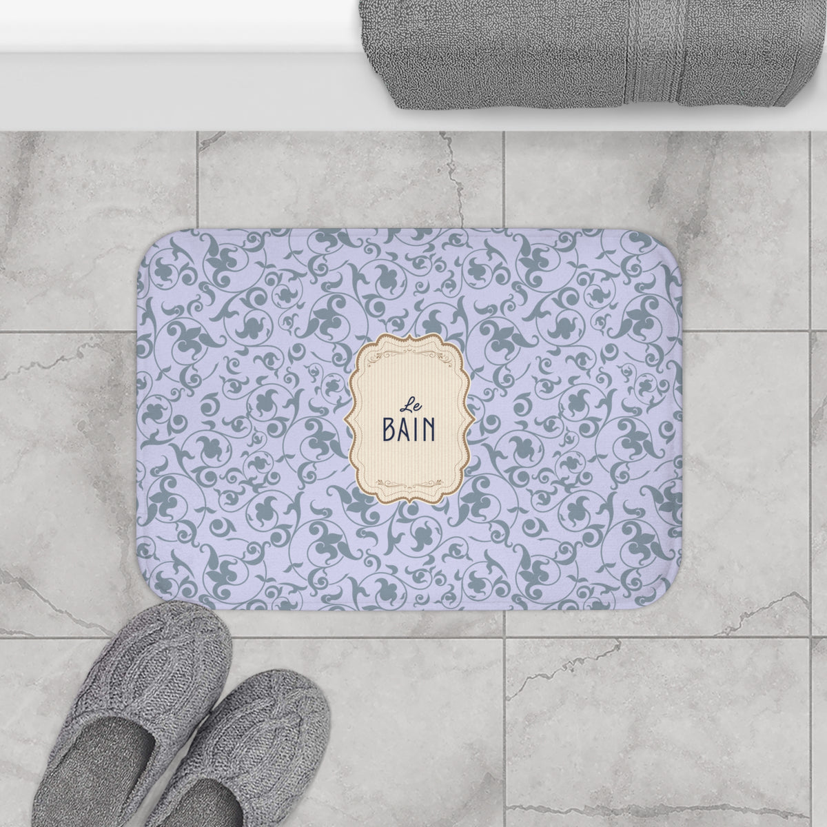 A rectangular bath mat with rounded corners on a light gray marble tile floor, featuring a lavender and gray floral scroll pattern with a central cream label that reads 'le BAIN'. Gray slippers and a folded gray towel are partially visible around it.