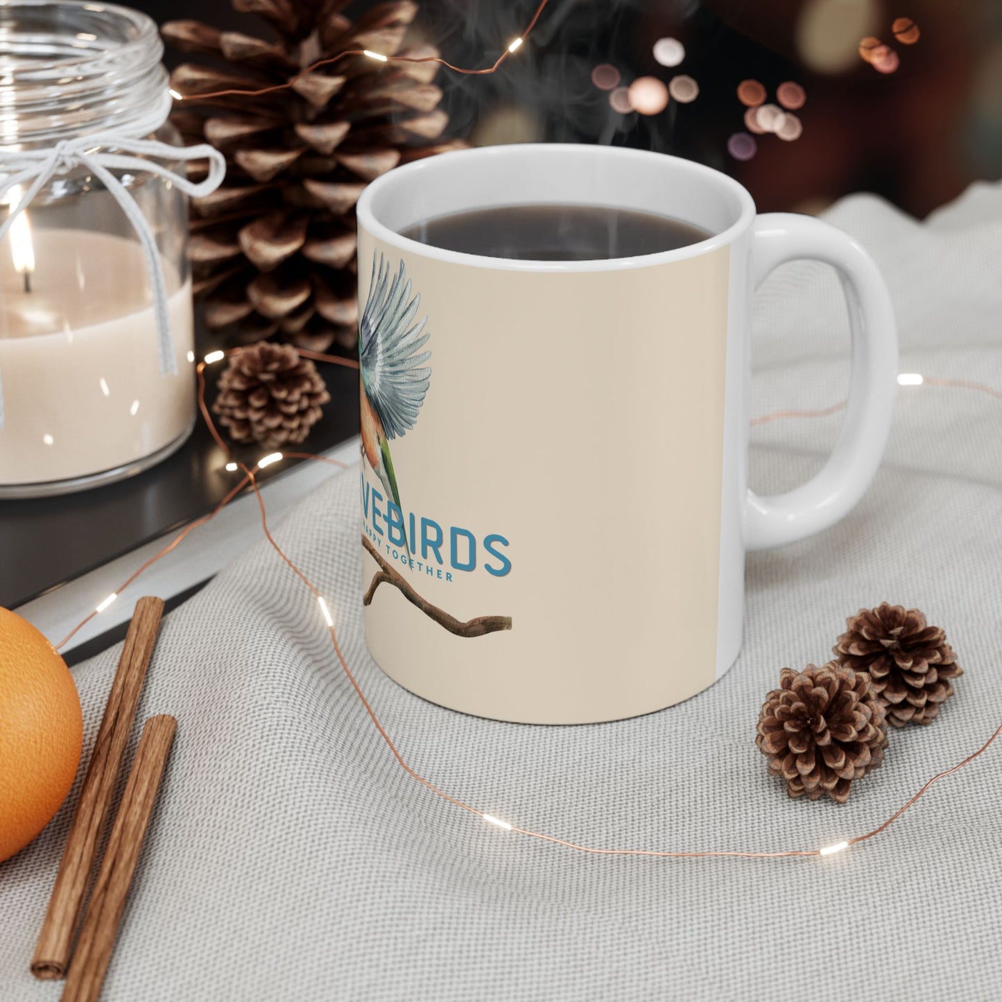 A white ceramic coffee mug with a cream-colored front panel, featuring a blue bird illustration and the text 'BLUEBIRDS CAFE'. The mug is filled with a hot beverage and styled on a light gray fabric tablecloth with pinecones, fairy lights, cinnamon sticks, an orange, and a glass jar candle in the background.