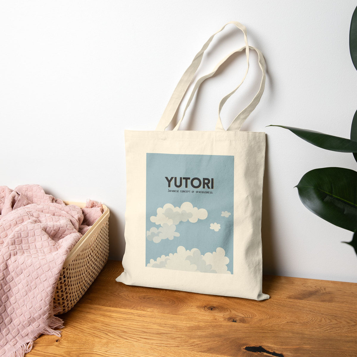 A cream canvas tote bag with long handles stands on a wooden surface against a white wall. The bag has a large square graphic print featuring a light blue sky and white clouds, with the word 'YUTORI' printed above.