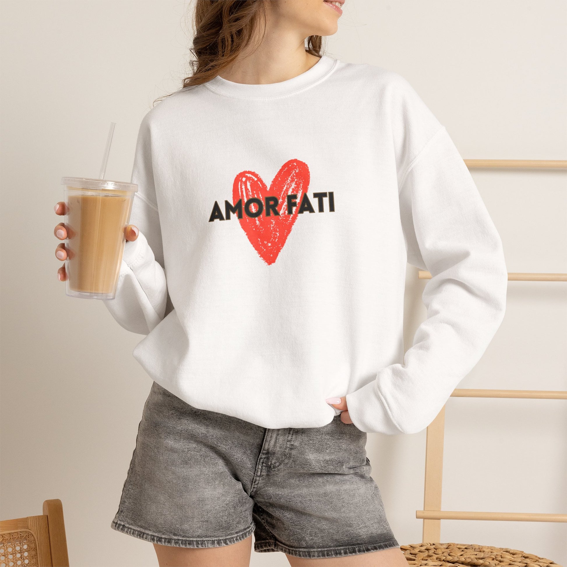 A woman wearing a white crewneck sweatshirt with a red heart and the black text 'AMOR FATI' printed on the front, paired with gray denim shorts and holding an iced coffee.