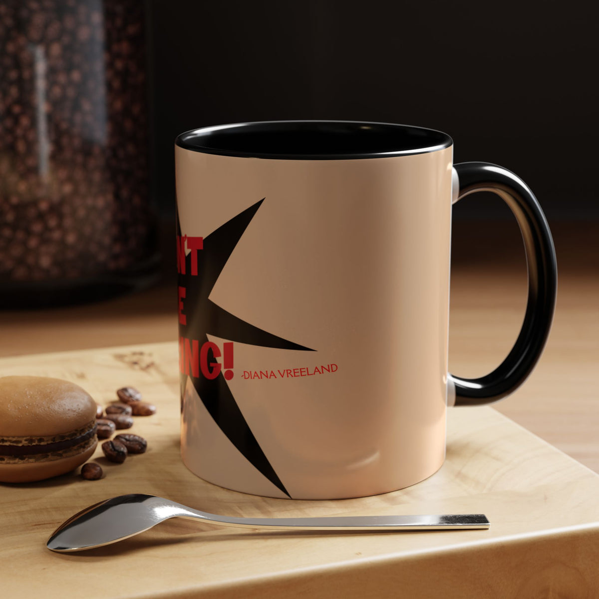 A ceramic coffee mug with a black handle and interior, featuring a beige exterior with a black starburst graphic and red text reading 'DON'T BE ENGIN! - DIANA FREELAND'. The mug sits on a wooden surface next to a spoon and coffee beans.